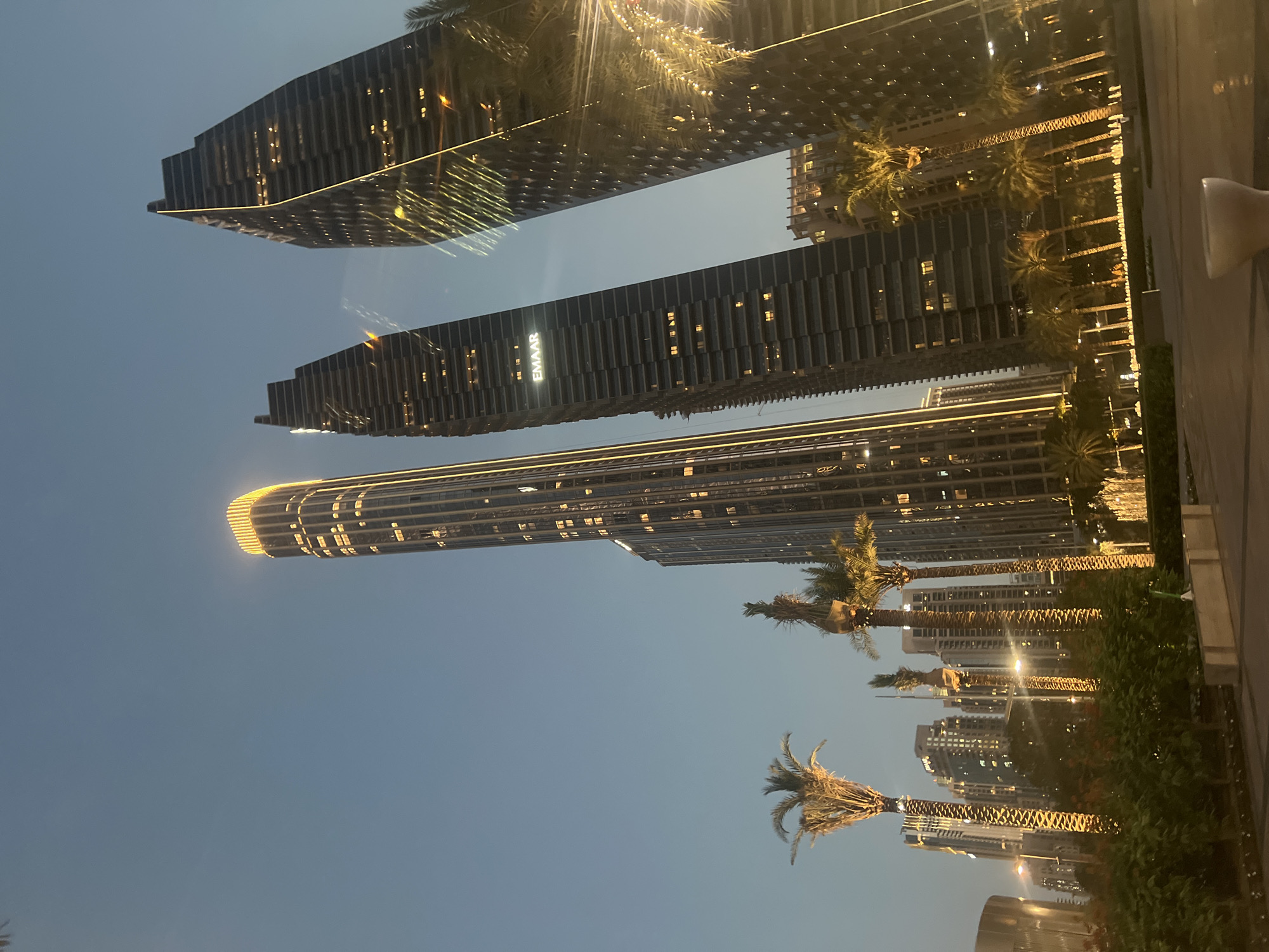 The provided image, oriented horizontally, depicts a vibrant evening or nighttime urban landscape in Dubai, UAE. Three modern skyscrapers extend across the left side of the frame, standing brightly illuminated against a clear, dark blue sky.
The topmost building in the frame features a stepped architectural design with a distinct horizontal golden light strip along its upper section. Below it, the middle skyscraper is rectangular and prominently displays the word "EMAAR" vertically in bright white letters on its facade. The third building, positioned at the bottom of the group, is notably slender and exceptionally tall, with its very top radiating a bright glow. Numerous windows on all buildings are lit, indicating activity within.
To the right, a meticulously maintained row of palm trees stretches horizontally, bordering what appears to be a paved pathway or street. Many of these palm trees are adorned with festive string lights, contributing to the luminous urban ambiance. Beyond the trees, areas of green foliage and manicured bushes are visible along the ground level. A small, white, cup-like object is partially visible on the far right edge of the frame. The scene captures a tranquil yet lively urban environment after sunset, with no specific human activity discernible.