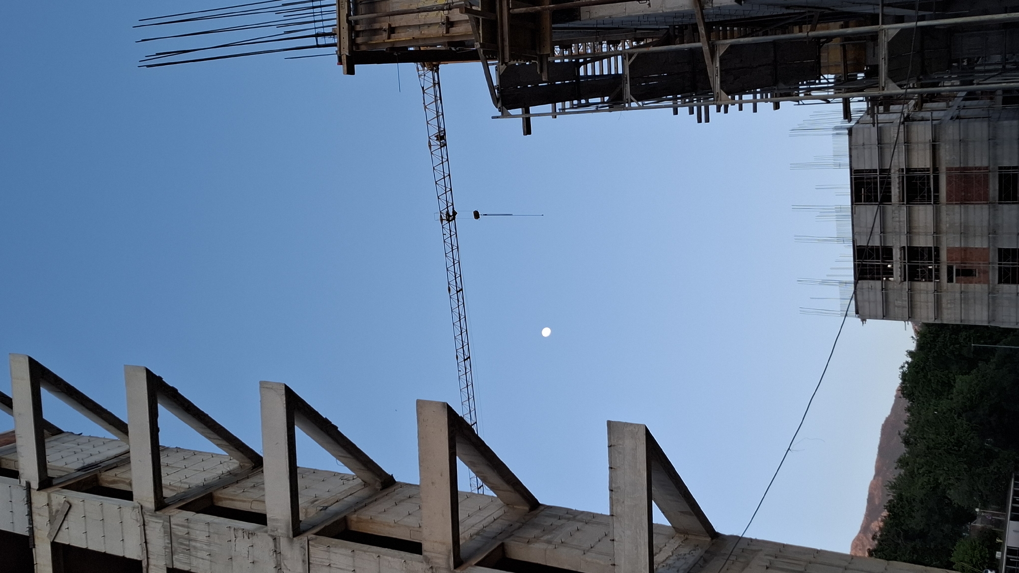 The image, oriented correctly, captures an upward perspective of a construction site under a clear, bright blue sky in Boreti, Montenegro. In the center of the sky, a distinct white, circular moon is visible, indicating daylight conditions.
A prominent construction crane dominates the left side of the frame, its long, lattice-like boom extending diagonally across the sky. Below it, in the lower left foreground, are concrete structural elements, possibly part of a roof or an upper floor, featuring exposed rebar or formwork arranged in a series of triangular shapes.
In the upper and right portions of the frame, parts of buildings under construction are visible. The top left shows exposed rebar protruding from an unfinished concrete structure. On the right, a multi-story building in progress features concrete walls, scaffolding, and numerous empty window openings, with more rebar extending upwards from its top.
Towards the bottom right, the verdant green foliage of trees and a section of a distant, rocky mountain range provide a natural backdrop to the man-made development. A thin, dark cable or wire runs diagonally across the middle-right of the sky, connecting various parts of the construction. The scene conveys a sense of ongoing development and progress. No people or discernible text are present in the image.