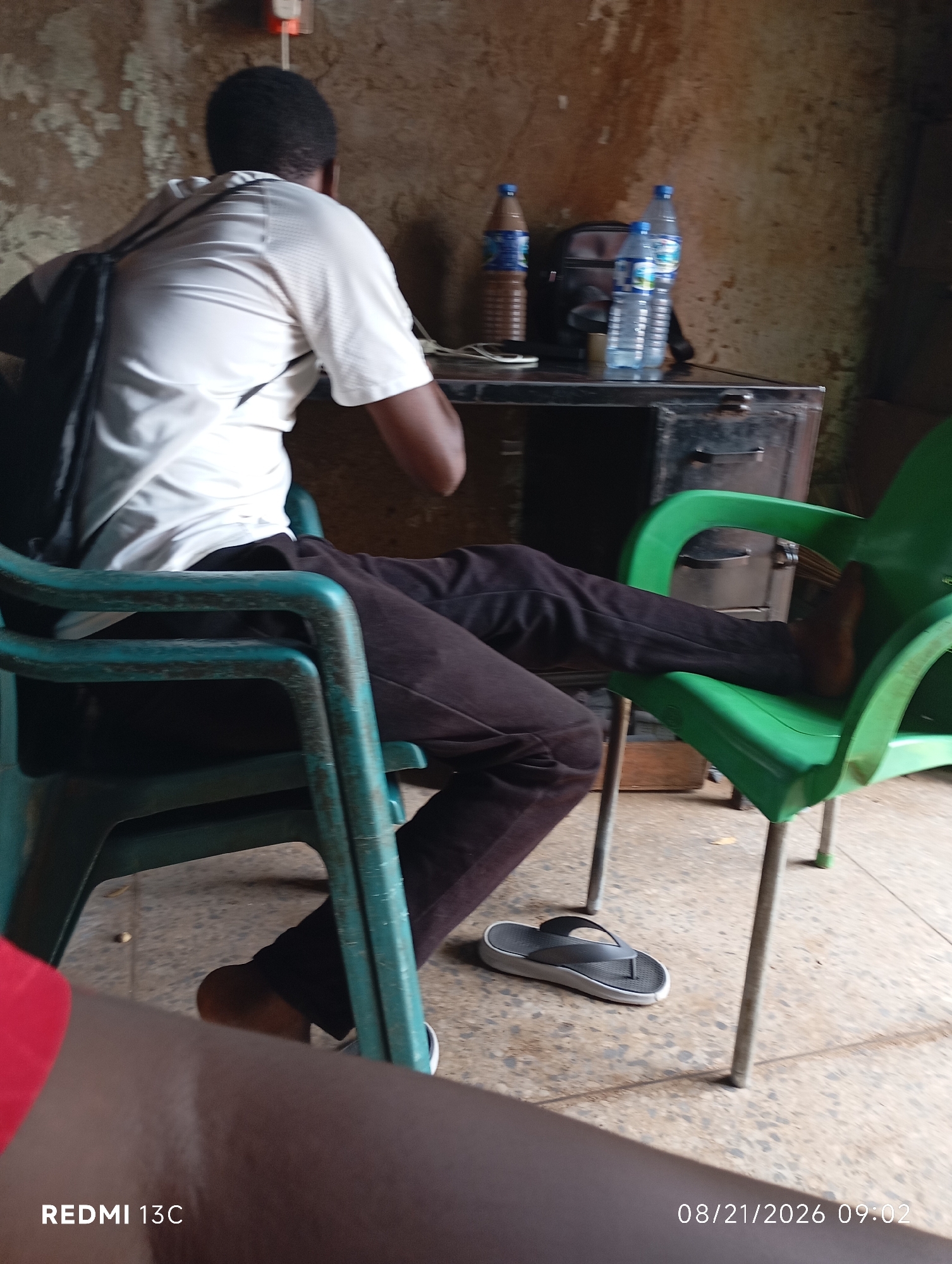 The image depicts a casual indoor scene, possibly a shop or office, in Shendam, Nigeria. A young Black male wearing a white t-shirt and dark trousers is seated in a teal plastic chair, facing away from the camera. He has a black backpack slung over his left shoulder. His right leg is extended, resting on a green plastic chair. A pair of dark flip-flops is visible on the speckled concrete floor to the left of the green chair. On a metal desk behind the seated individual, two bottles of water and a bottle of what appears to be chocolate milk are placed next to a dark bag. The wall behind the desk is textured and appears to be a light brown color. The overall lighting suggests an indoor environment with ambient light. The timestamp "08/21/2026 09:02" is visible in the bottom right corner of the image, along with "REDMI 13C" in the bottom left.