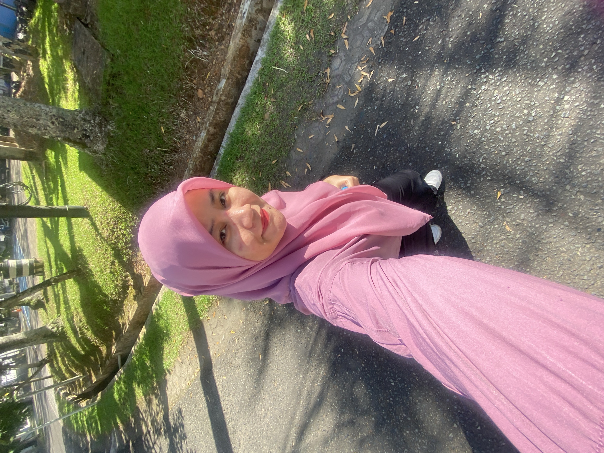 A young woman wearing a pink hijab and a pink long-sleeved shirt is captured in a high-angle selfie outdoors. She is standing on an asphalt path, possibly in a park or recreational area in Banjarbaru, Indonesia, given the presence of trees, grass, and a drainage ditch. Sunlight casts shadows on the ground, indicating it is daytime. The woman's face is partially visible, and she appears to be smiling. She is wearing black pants and white sneakers. The background features a well-maintained grassy area with scattered leaves, and several trees. The overall impression is a casual, sunny day.