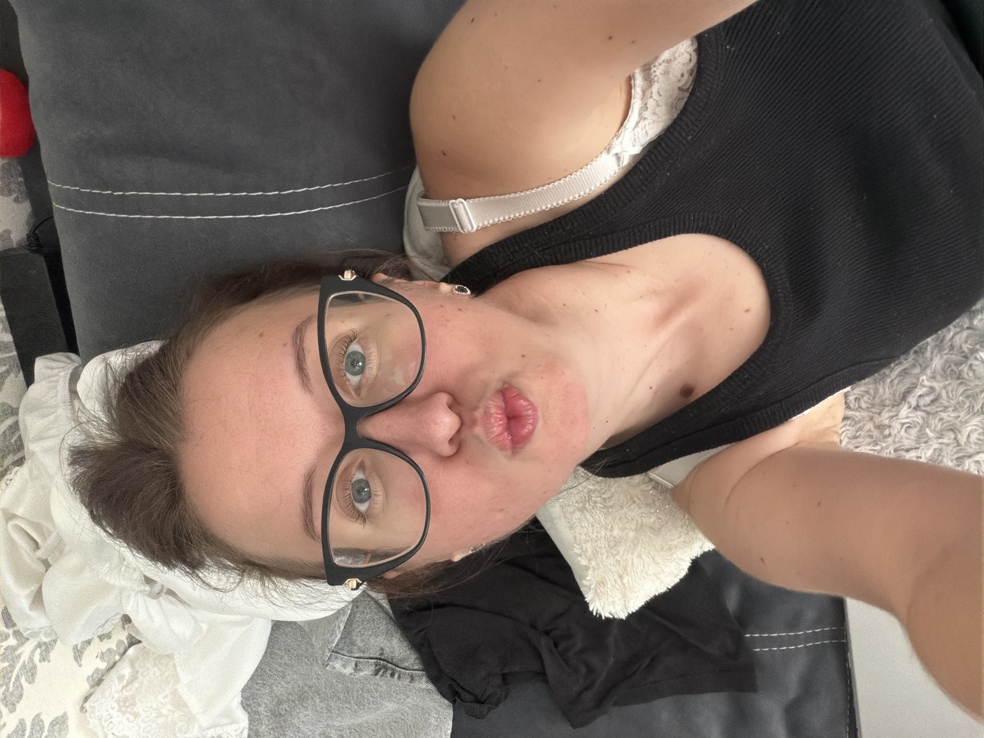 In this rotated, close-up selfie, a woman with blue eyes and dark hair, wearing black-rimmed glasses, is lying down, possibly on a couch, with her face tilted towards the camera. She is wearing a black tank top with a white lacy bra underneath. Her lips are pursed in a kissing gesture. The background includes a dark gray textured surface, possibly a sofa, with white stitching. There are also some white and gray fabrics visible, suggesting laundry or bedding. The lighting appears to be indoor, possibly natural light. The overall mood is casual and playful. There is no visible text in the image. The location is identified as Bila Tserkva, Ukraine, but there are no specific visual cues in the image to confirm this.