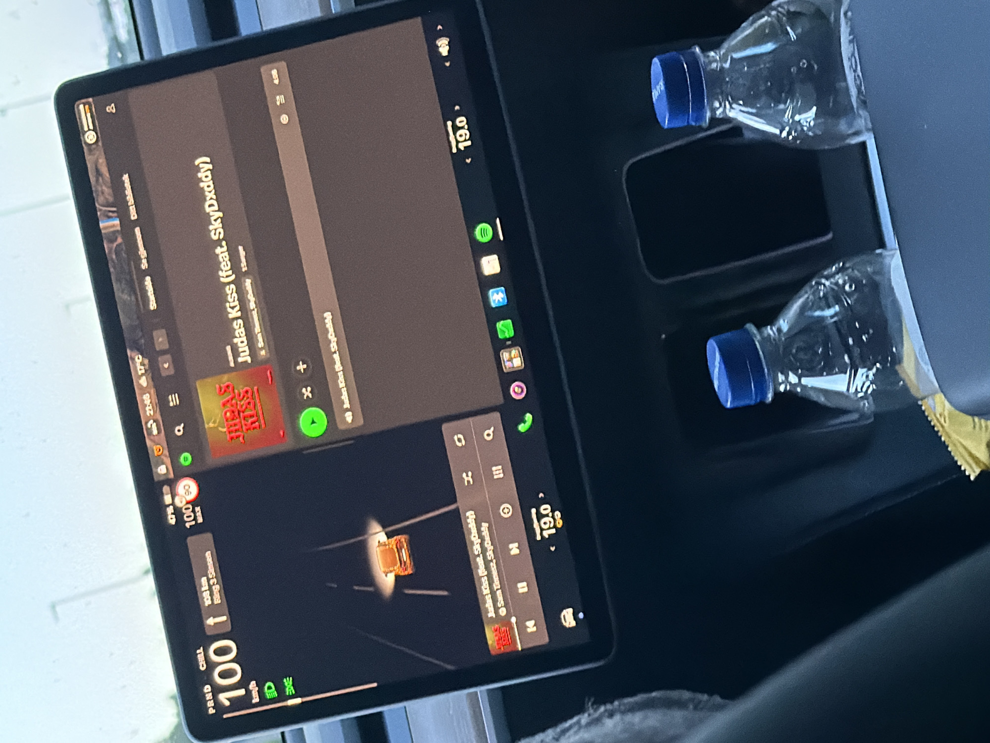 The image shows the interior of a car, likely a Tesla, with its large central touchscreen display prominently in focus. The screen displays navigation information, music playback, and car controls. The car is in motion, indicated by the navigation view showing a car icon on a road. The speed displayed is 100 km/h. The time shown on the screen is 21:46 and the temperature is 17°C. Music is playing, identified as "Judas Kiss (feat. SkyDxddy)". Two plastic water bottles with blue caps are visible in the center console. The overall environment suggests a daytime scene, possibly overcast, given the lighting. The location is identified as Sponvika, Norway.