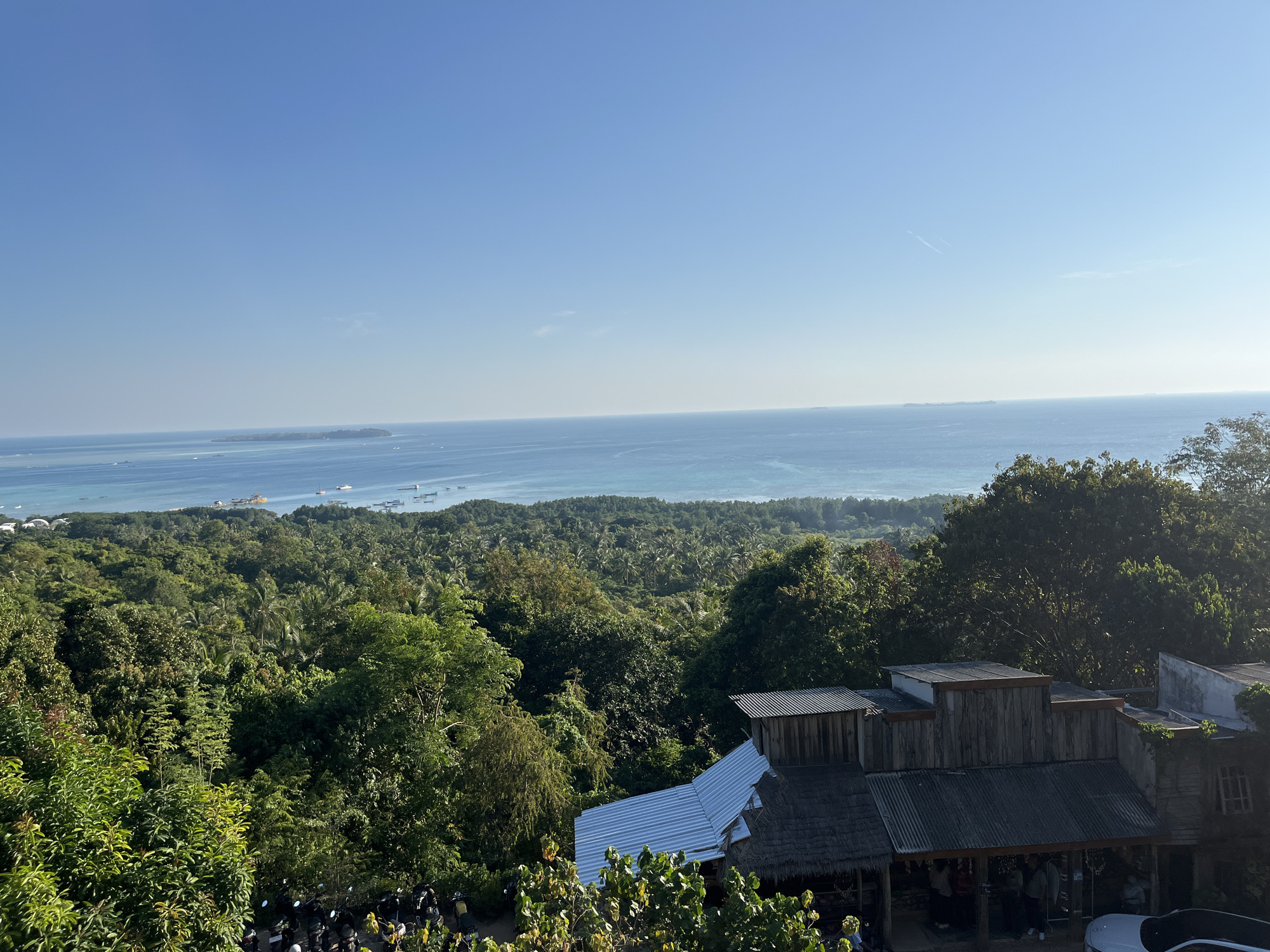 The image shows a panoramic view of the city of Karimunjawa, Indonesia, on a clear day. The foreground features a dense green forest with a few buildings nestled amongst the trees. The buildings have a rustic appearance with corrugated metal roofs and wooden walls. A row of motorcycles is parked in the lower left corner of the image. Beyond the forest, a vast expanse of blue ocean stretches to the horizon, dotted with several boats and a distant island. The sky is a clear, bright blue, with a few faint white clouds. The scene suggests a tropical, coastal environment, likely a popular tourist destination due to the presence of boats and the scenic view.
