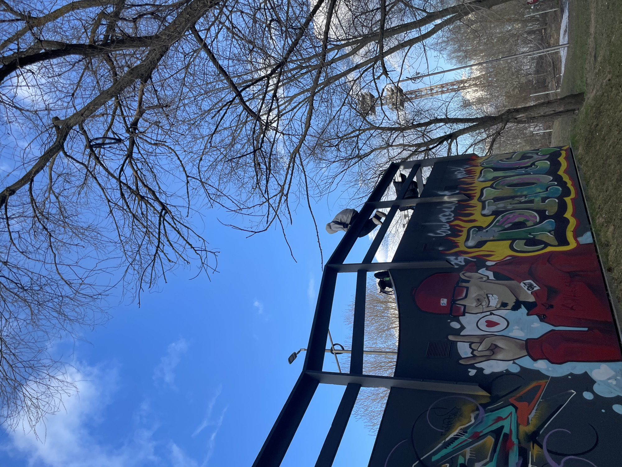This image, taken in Izhevsk, Russia, captures an upward-looking perspective of an outdoor scene under a bright blue sky with scattered white clouds.
The primary subject is a large, dark, multi-level metal structure, possibly part of a skate park, parkour course, or urban art installation. Its side is vibrantly decorated with graffiti. A prominent section of the graffiti features a cartoonish male character with light skin, wearing a red backward baseball cap, glasses, and a red jacket. He sports a beard, is smiling broadly, and is making a "rock on" hand gesture with his right hand, with a heart symbol nearby. Above him, stylized letters are visible, possibly "IZH" or "SHOW," surrounded by flame-like designs. Other abstract graffiti elements adorn the lower parts of the structure.
Two individuals are seen on top of or interacting with this structure. One person, dressed in white, is visible near the upper edge, seemingly climbing or perched. Another figure is partially discernible further back on the structure. Their presence suggests recreational activity, possibly involving urban sports or exploration of the structure.
Bare, deciduous tree branches fill the upper and left portions of the frame, silhouetted against the sky, indicating late autumn, winter, or early spring. In the distance, beyond the trees, a tall metal structure resembling an amusement park ride or roller coaster is visible. The ground area to the right shows grass, suggesting a park setting.
The bright, even lighting points to a clear day, likely midday or early afternoon, with cool weather. The visible graffiti adds a distinct urban art character to the location.