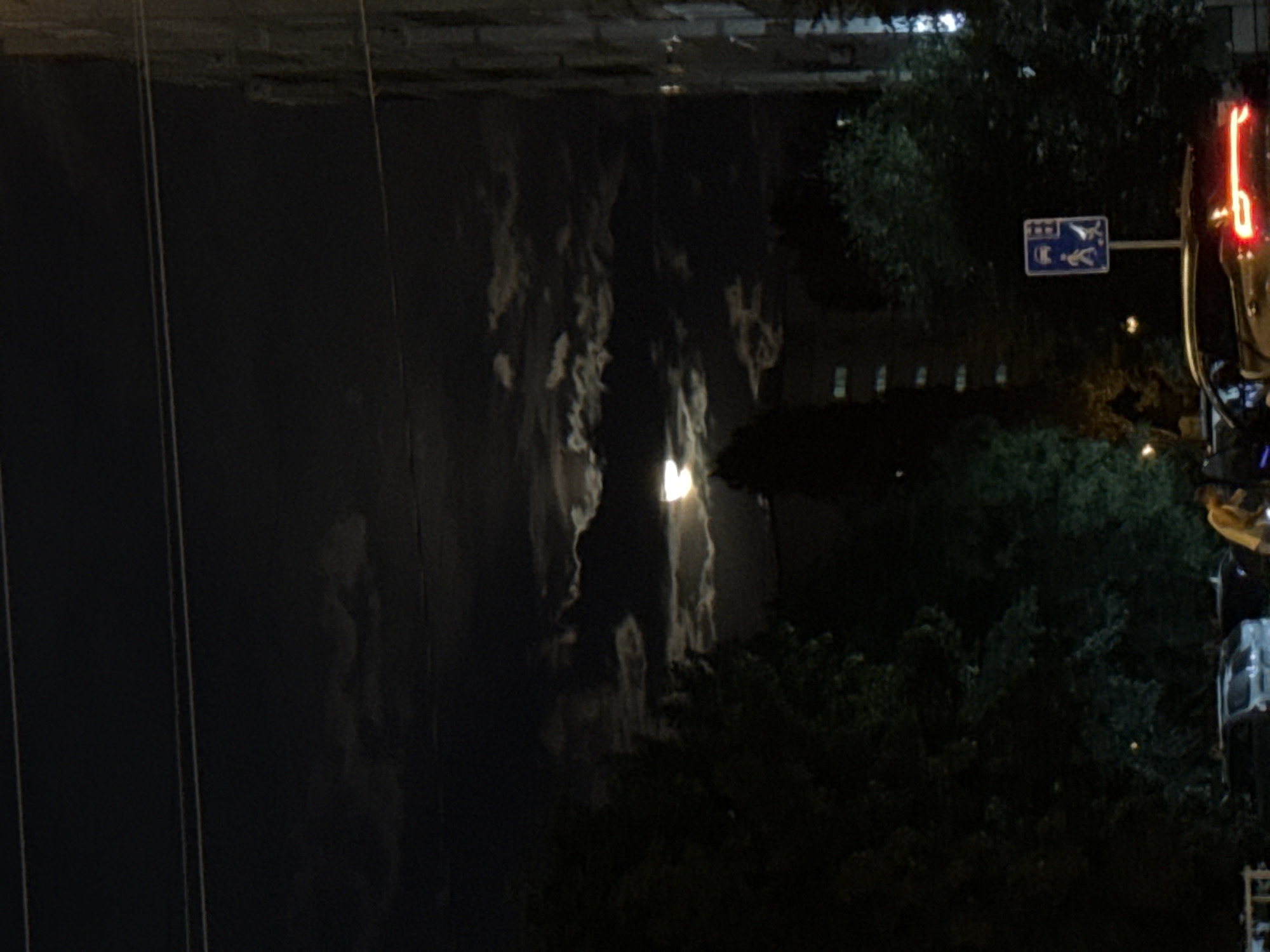 A nighttime scene in Tolyatti, Russia, features a nearly full moon partially obscured by clouds, casting a dim light over the urban landscape. The moon is positioned slightly to the left of the center of the frame, amidst swirling dark clouds with illuminated edges. In the foreground, dark trees line the street, their foliage dense and silhouetted against the night sky. To the right, a multi-story building stands, its windows dark. A street sign indicating pedestrian and vehicle passage is visible near the center-right, with a car parked partially in front of it. The car has its taillights illuminated, casting a red glow. A person in a yellow shirt is visible standing next to another vehicle, possibly interacting with it. Several other cars are parked on the street. Overhead, power lines stretch across the dark sky. The overall atmosphere is one of quiet evening activity in a residential or mixed-use urban area.