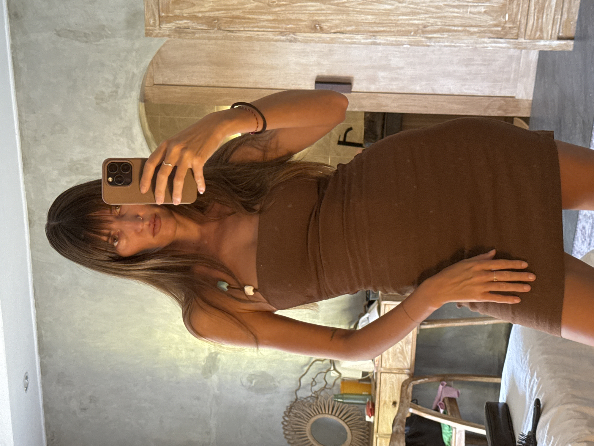 A woman is taking a mirror selfie in a room. She is wearing a strapless brown dress and has long brown hair with bangs. She is holding up a smartphone in her right hand. Her left hand is resting on her hip. In the background, there is a wooden dresser with a mirror on it, a beaded necklace hanging from her shoulder, and a large wooden door. The room appears to be a bedroom or dressing area. The overall lighting is natural and diffused. This appears to be taken in a hotel room in North Kuta, Indonesia.