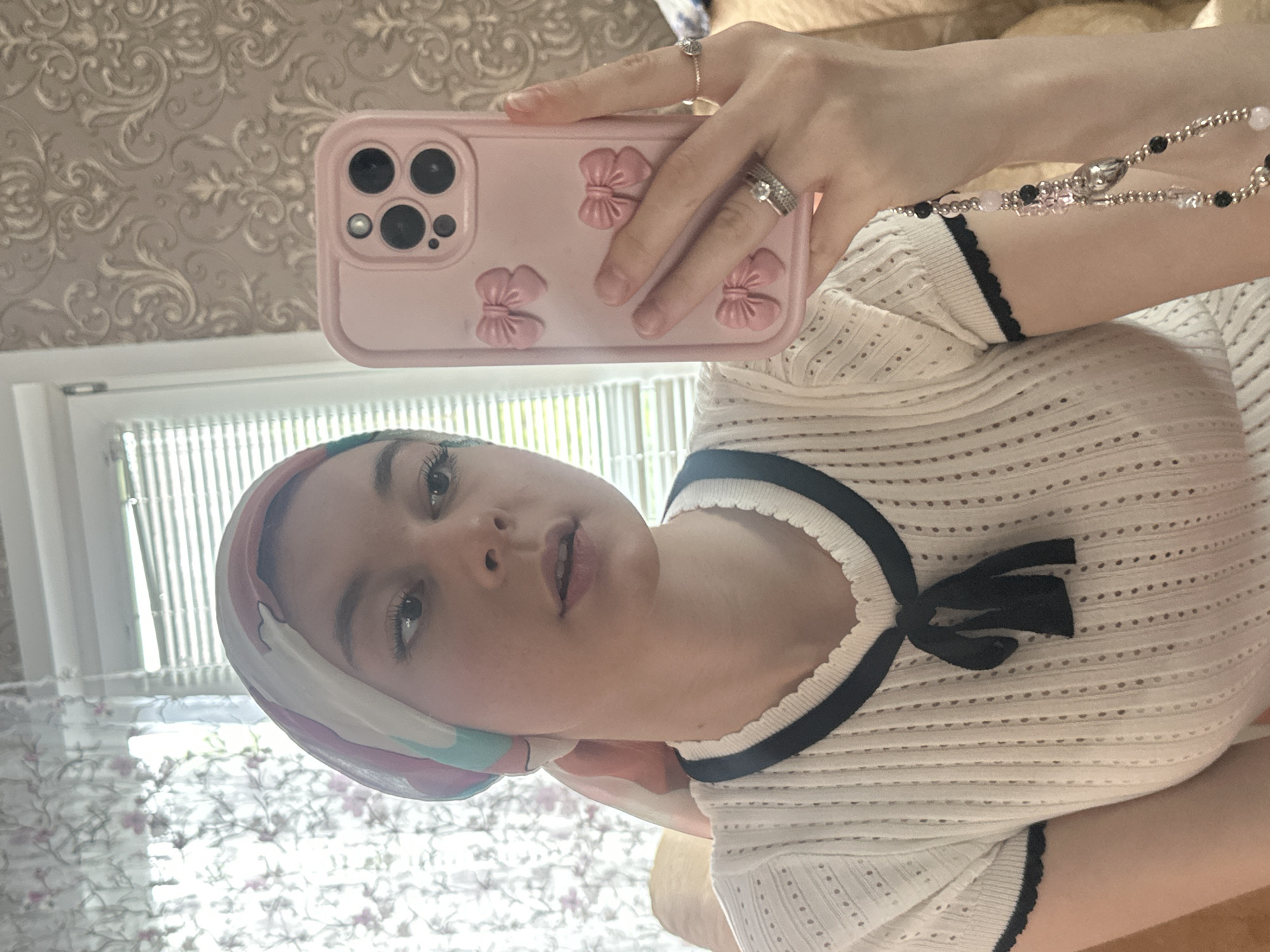 A young woman is taking a selfie in a mirror. She is wearing a patterned headscarf and a white top with a black trim and bow at the neckline. She holds a pink cell phone case with bow decorations. She is wearing rings. The background reflects a window with blinds and patterned wallpaper. There is no information to suggest this photo was taken in the city of Коноваловка, Russia.