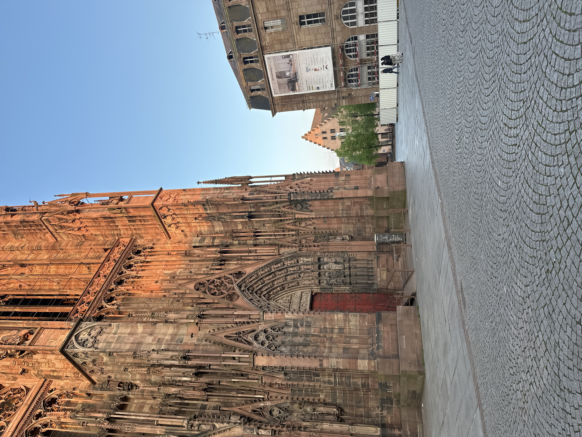 The image shows the Cathédrale Notre-Dame de Strasbourg, a prominent landmark in Strasbourg, France. The cathedral's facade is richly decorated with Gothic architectural details. The building is constructed from red sandstone. To the right of the cathedral, there is a paved area, likely a pedestrian zone. In the background, additional buildings and a blue sky can be observed. The image is taken at a low angle, emphasizing the height of the cathedral. There are some people. There are also barriers present.