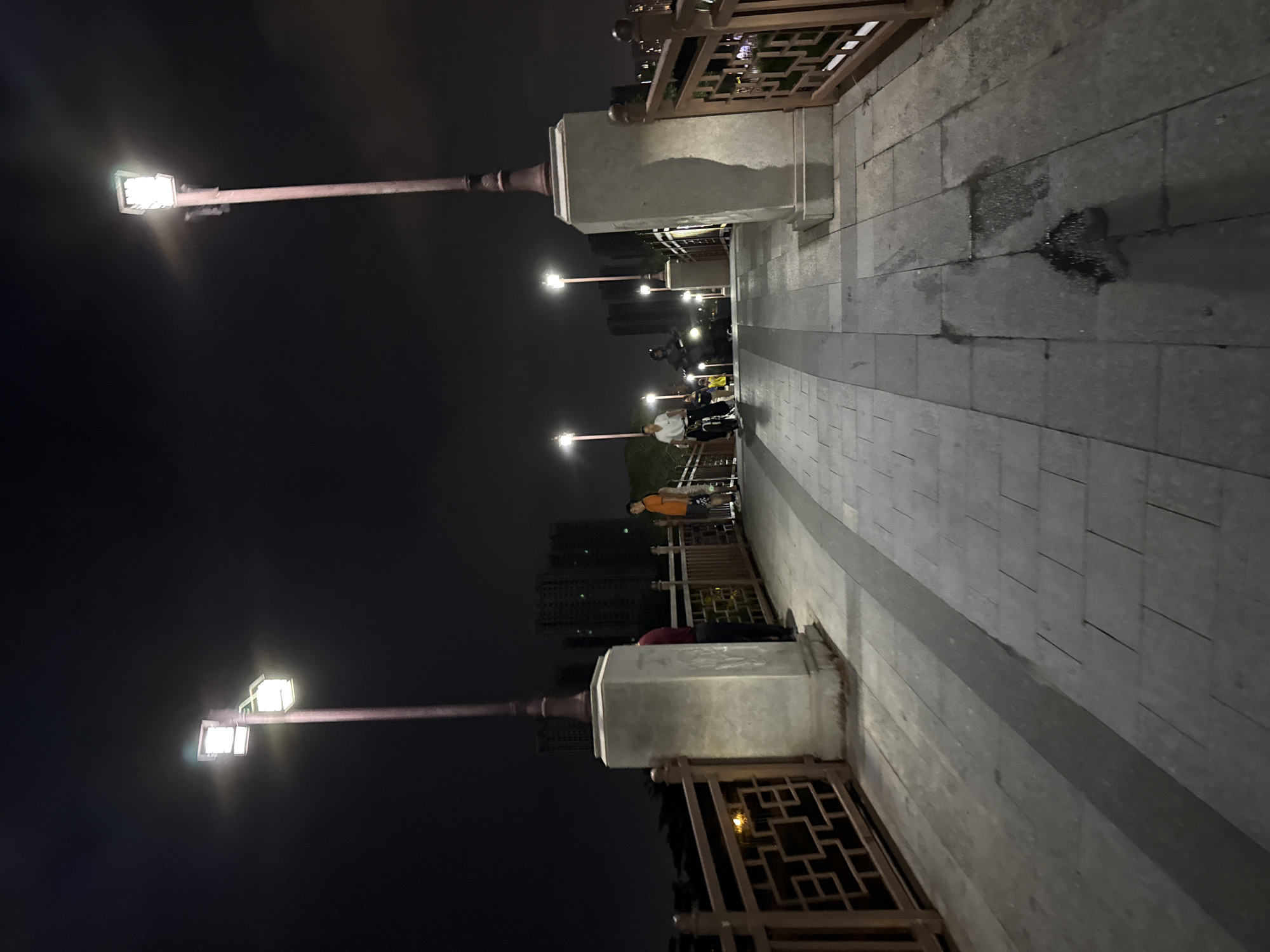 The image depicts a nighttime scene on a pedestrian bridge in Huizhou, China. Two ornate lampposts with modern LED fixtures illuminate the walkway, which is paved with grey stone tiles. The bridge has decorative wooden railings with a lattice pattern. In the background, several tall apartment buildings with illuminated windows are visible against the dark sky.
Several people are present on the bridge. On the left, two individuals are standing near a lamppost and a concrete pedestal. Further down the bridge, a person is riding a bicycle, and another person is riding a motorcycle. Another individual in an orange shirt stands on the right side of the bridge, looking away from the camera. The ground on the bridge appears slightly wet in places, suggesting recent rain or cleaning. The overall atmosphere is calm and urban, characteristic of a city at night.
