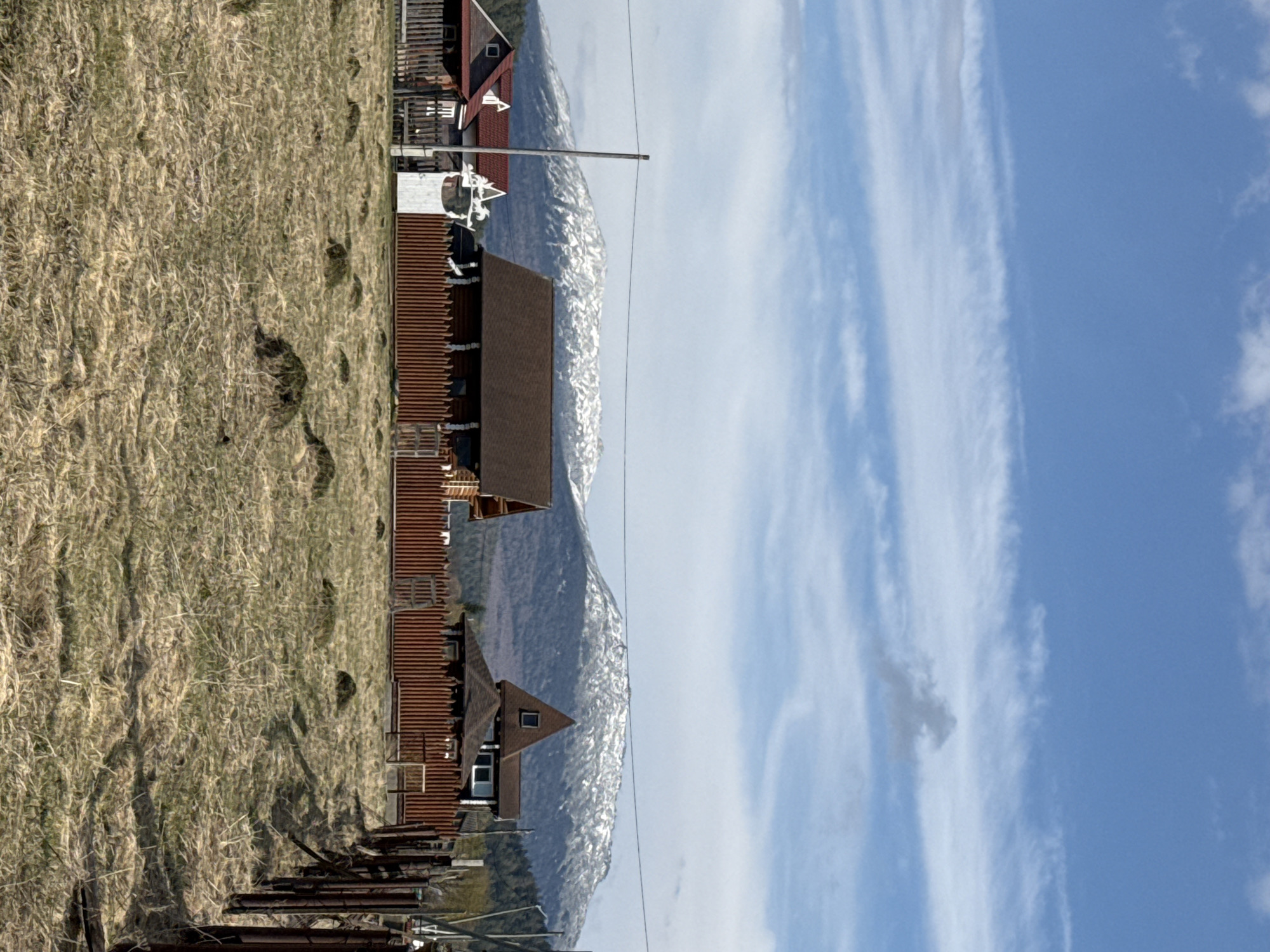 The image depicts a rural scene in Tyulyuk, Russia. In the foreground is a field of dry, yellowed grass. Adjacent to the field are several wooden structures, including residential houses with red roofs. In the background, snow-capped mountains rise into a blue sky with streaks of white clouds. A power line is visible stretching horizontally across the image. There are no people visible in the image.