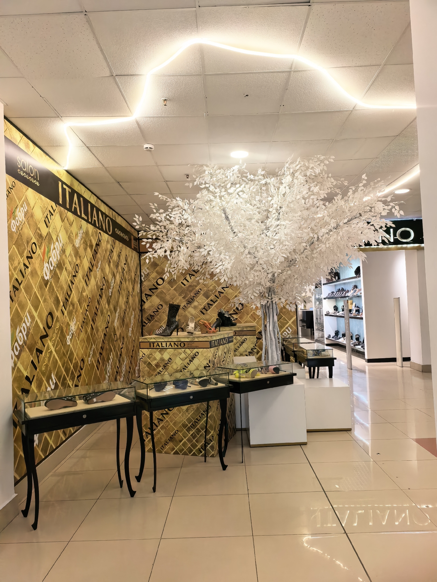 An interior shot captures a store display with a prominent, artificial white tree as a central decorative element. The store's wall features a repeating pattern of the word "ITALIANO" in large black lettering against a textured gold background. Several display cases showcase various footwear and accessories. In the background, a shelf display reveals more shoes. A white ceiling with square panels is illuminated by recessed lighting and a wavy neon light strip. The floor is tiled in a light beige color. The overall impression is of a retail environment, likely a shoe or fashion boutique. The image is taken from a slightly low angle, looking towards the back of the store.