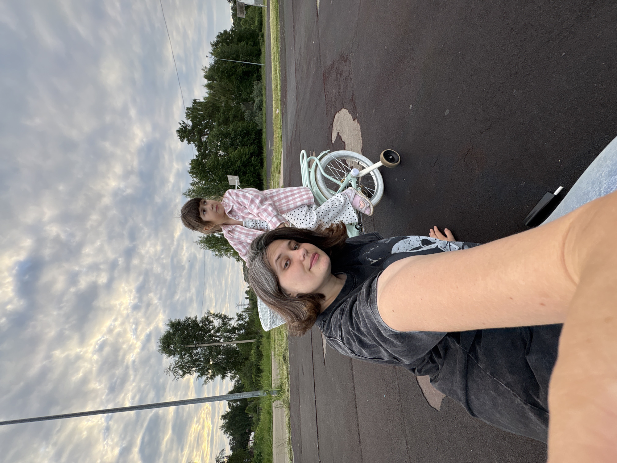 In Chaikovsky, Russia, on a paved outdoor area with scattered dark patches, a woman is in the foreground, taking a selfie. She has medium-length dark hair with some grey streaks, and is wearing a dark grey t-shirt with a graphic print and denim jeans. Behind her, a young girl with brown hair is sitting on a light blue bicycle with training wheels. The girl is wearing a pink and white checkered shirt over a white polka dot outfit. The background consists of trees, a cloudy sky, and what appears to be a sports court or play area with a basketball hoop visible.