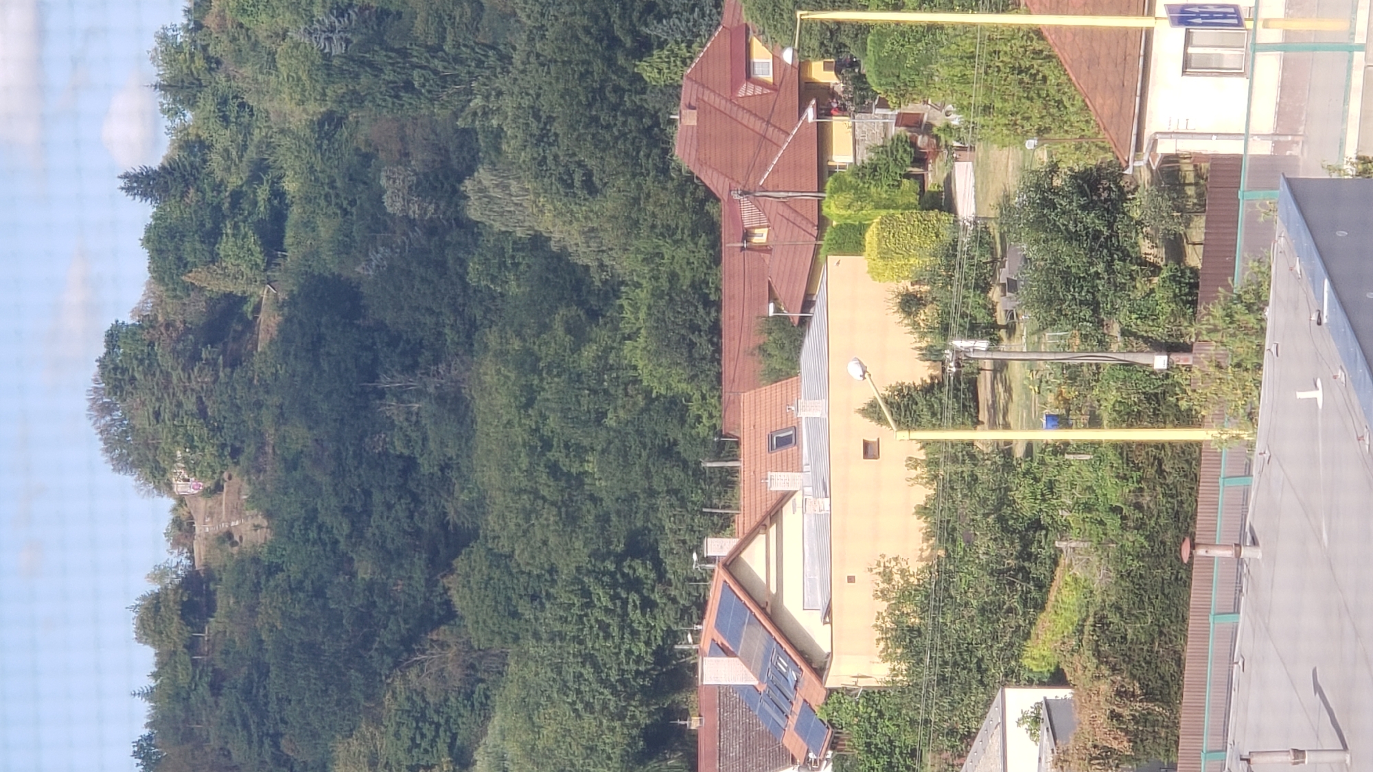 This is a photograph taken from a high vantage point, looking down on a residential area surrounded by dense trees. In the foreground, the roof of a building is visible, with several ventilation pipes and a partial view of a building's wall. Behind this, a row of houses with red-tiled roofs can be seen, some with solar panels installed on their slopes. The houses are a mix of yellow and light-colored facades. Beyond the houses, a steep, tree-covered hill dominates the background. A path with steps is visible winding up the hill, leading to a structure at the summit. The overall impression is of a suburban or semi-rural setting with a prominent natural landscape feature.