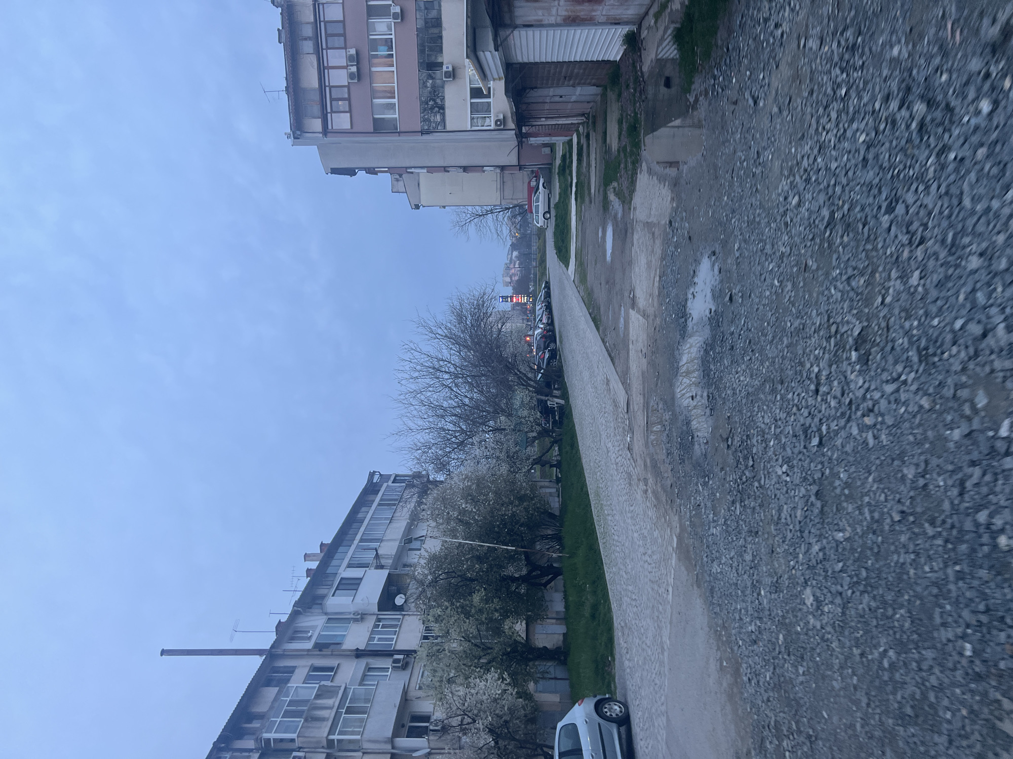 The image shows a street in Yambol, Bulgaria. The sky is overcast. Buildings stand on either side of a narrow sidewalk, which transitions to a gravel path. Several parked cars are visible along the street. Trees are present along the sidewalk, some with white blossoms. The ground appears damp, with puddles on the gravel path. There are no people visible in the frame.