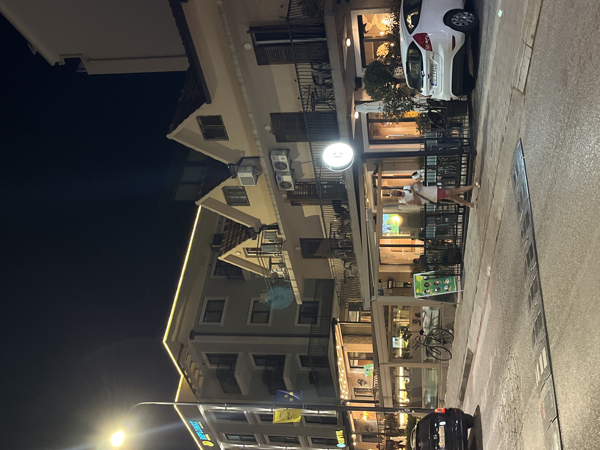 An exterior night shot of a street scene in Budva, Montenegro, shows a multi-story building with a prominent "DIPLOMAT HOTEL" sign. The building has balconies with railings, and some windows are illuminated. A streetlamp is visible on the left, casting light on the building and the sidewalk. Several people are walking or standing on the sidewalk, and there are outdoor seating areas with tables and chairs associated with ground-floor establishments. A black car is parked on the street on the left, and a white car is parked on the right. A bicycle is leaning against a railing on the sidewalk. A speed bump is present on the asphalt road in the foreground.