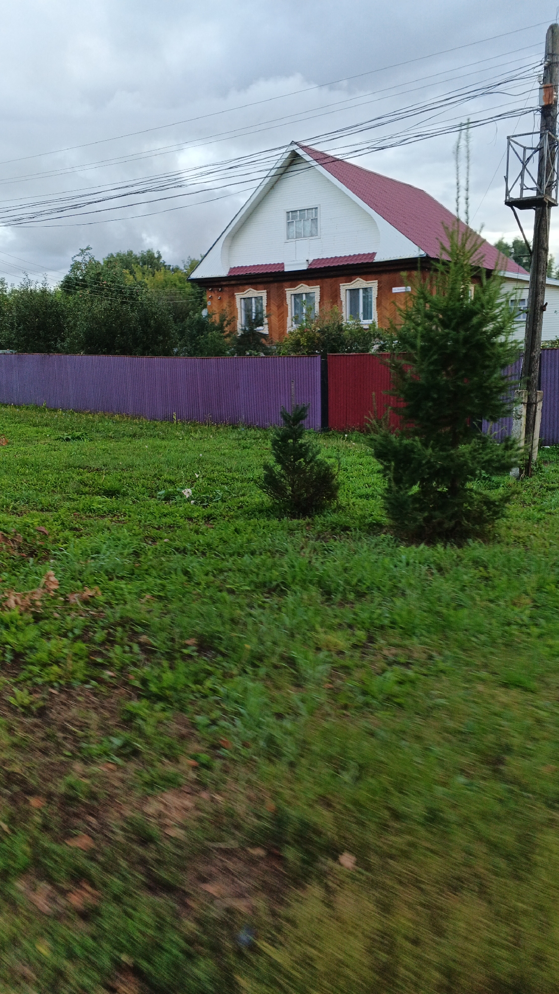 In the village of Podgornoye, Russia, a wooden house with an orange facade and a red roof is visible behind a vibrant purple corrugated metal fence. The house has white trim around its windows, and the upper gable is also white with a window. Two small evergreen trees stand in the grassy yard in front of the fence. A wooden utility pole with electrical wires runs along the right side of the frame. The sky is overcast with gray clouds. The foreground is blurred, indicating motion, likely from a vehicle.