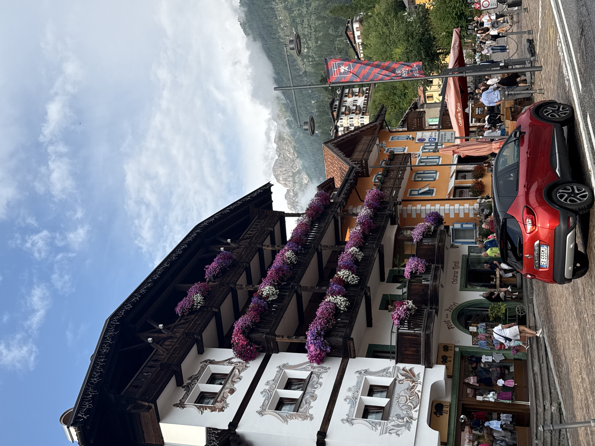 In the city of Moena, Italy, a red Renault Captur is parked on a cobblestone street in front of a building featuring alpine-style architecture. The building has multiple balconies overflowing with vibrant purple and pink flowers. The ground floor of the building houses businesses, including a shop selling clothing and a restaurant called "Osteria Tirol". Several people are visible walking along the sidewalk and seated at outdoor tables. In the background, a mountainous landscape with lush green trees and a cloudy sky can be seen. A tall flagpole with a red and black banner stands on the right side of the street.
