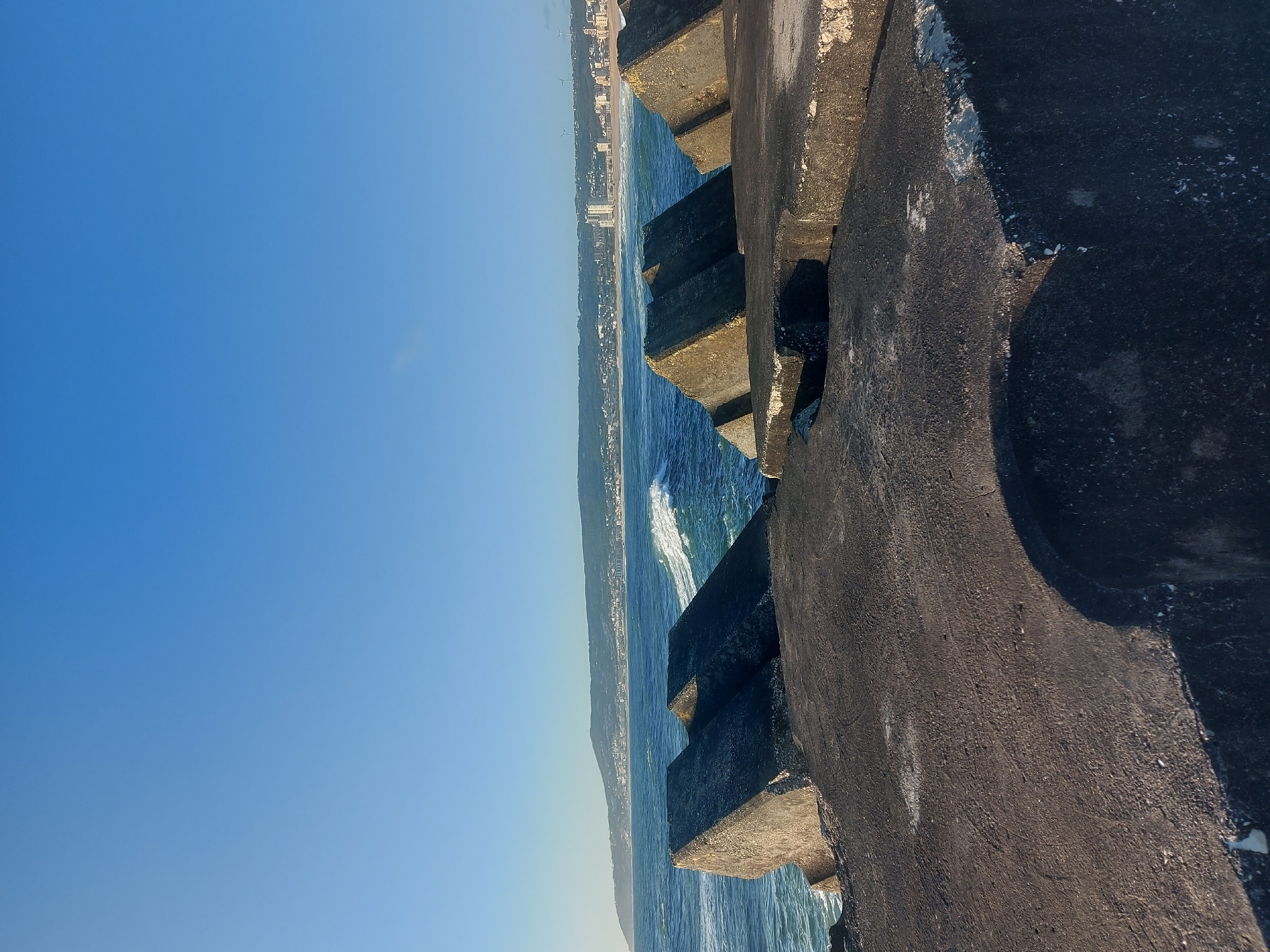This image captures a vibrant coastal scene in Figueira da Foz, Portugal, under a clear blue sky. The perspective is from a large concrete structure, likely a pier or breakwater, extending into the ocean.
In the foreground, dominating the right side of the frame, are massive, dark grey concrete blocks. These structures, possibly tetrapods or similar coastal defense elements, exhibit rough textures and lighter patches, suggesting wear from the sea and elements. They appear sturdy and functional, forming a barrier against the waves.
To the left of these concrete blocks, the deep blue waters of the Atlantic Ocean are visible. Gentle waves crash against the structures, creating streaks of white foam that contrast with the dark water. The water appears clear and bright under the sunlight.
Stretching across the horizon in the distance is a long coastline. Along this shore, a city or town is visible, characterized by numerous buildings lining the beach. Behind the urban area, rolling hills or mountains rise, covered in what appears to be lush greenery, adding depth to the landscape.
The sky is a vast and cloudless expanse of bright, uniform blue, indicating excellent weather conditions. The strong sunlight illuminates the entire scene, casting sharp shadows on the concrete blocks and highlighting the textures of the land and sea.
No people or identifiable activities are taking place, creating a serene and expansive atmosphere. The time of day appears to be mid-morning or early afternoon due to the bright, high sun. No visible text is present in the image. The scene beautifully blends man-made coastal defenses with the natural beauty of the ocean and distant shoreline.
