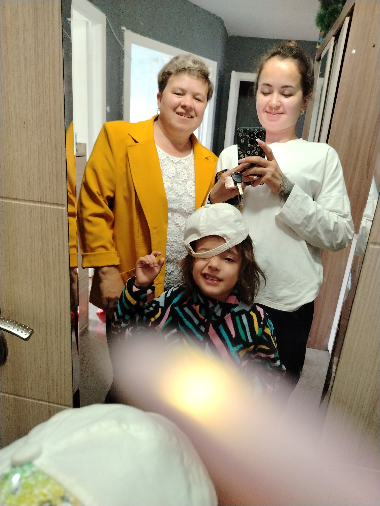 A selfie is taken in what appears to be a domestic setting, possibly an apartment in Noviy, Russia. Three individuals are visible in the mirror reflection. On the left, an adult with short, gray hair wears a white lace top and a vibrant yellow blazer. To her right, another adult, with dark hair pulled back in a bun, is holding a smartphone with a floral pattern case, taking the photo. She is wearing a white long-sleeved shirt and black pants. In the foreground, a child with long, brown hair and a white baseball cap pulled low over one eye is smiling. The child wears a colorful striped jacket. The background shows a gray wall, a wooden door frame, and part of a wooden wardrobe. The lighting suggests an indoor environment.