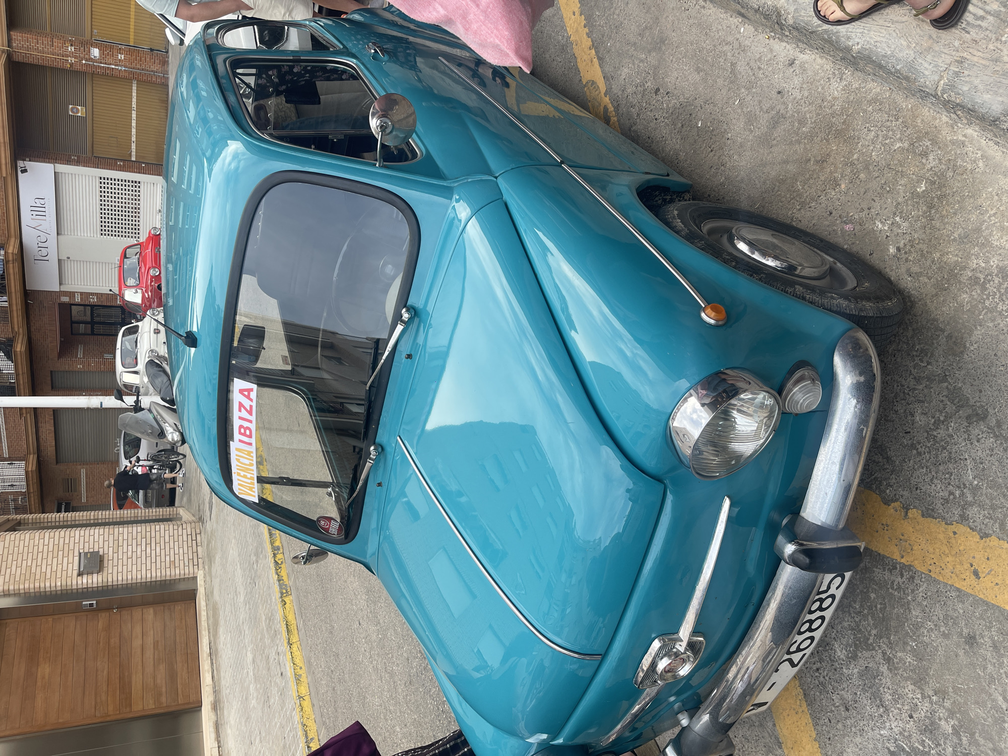 The image depicts an urban street scene in Manises, Spain, featuring a prominently displayed turquoise classic car.
In the foreground and center, a vintage turquoise car, identified by its "V-26885" license plate as registered in Valencia, is parked on a concrete surface marked with faded yellow lines. A white sign with orange text "VALENCIA IBIZA" is affixed to its front windshield, along with a smaller red circular sticker bearing "SEAT 600" text. The car features a chrome front bumper, circular headlights, amber turn signals, and a chrome trim strip running along the hood and sides. The hood reflects a building facade and overcast sky.
In the mid-ground, behind the turquoise car, two other classic cars, both appearing to be similar models (likely SEAT 600s), are parked: a red one partially visible behind the turquoise car's roof, and a white one further back, partially obscured by a grey scooter. A person wearing a dark t-shirt and patterned shorts is visible walking a black bicycle on the sidewalk to the left of the scooter.
The background consists of several buildings. To the left, a modern building features a large wooden panel door and a brick facade with a mailbox. To the right, a storefront with a white roller shutter displays a sign reading "Tere/Milla" and smaller text below it. Adjacent to this, another building has a brown garage door.
Partial views of individuals are present: the lower part of a person's leg clad in dark purple and a patterned garment is visible on the far left, and a bare foot wearing a dark sandal is visible on the concrete on the far right. A piece of pink fabric is also visible near the right side of the turquoise car. The ground is a textured concrete surface. The scene is illuminated by ambient light, suggesting an overcast day.