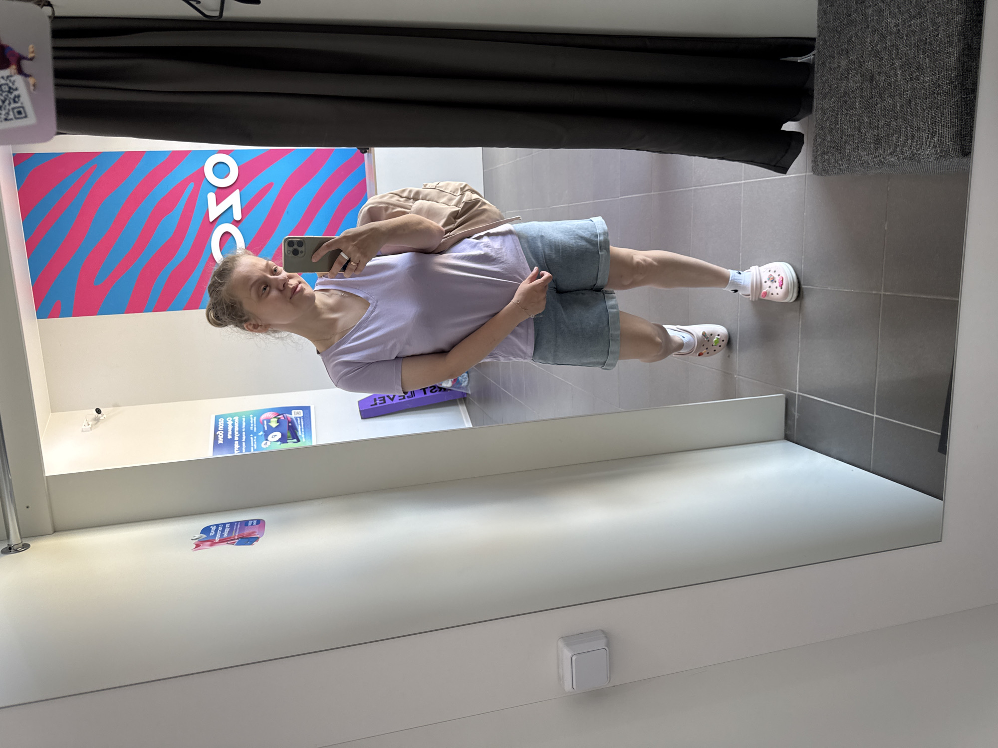A young woman is taking a mirror selfie in a fitting room, located in the city of Vyazniki, Russia. She is wearing a light purple v-neck t-shirt, denim shorts, white socks, and pink Crocs. She is holding a smartphone in her right hand and a light brown backpack is slung over her left shoulder. The fitting room has white walls and grey tiled flooring. In the background, there is a large poster with a pink and blue wavy pattern and the word "OSO" in white letters. A dark grey curtain hangs to the right of the woman, and a grey upholstered bench is visible in the bottom right corner.