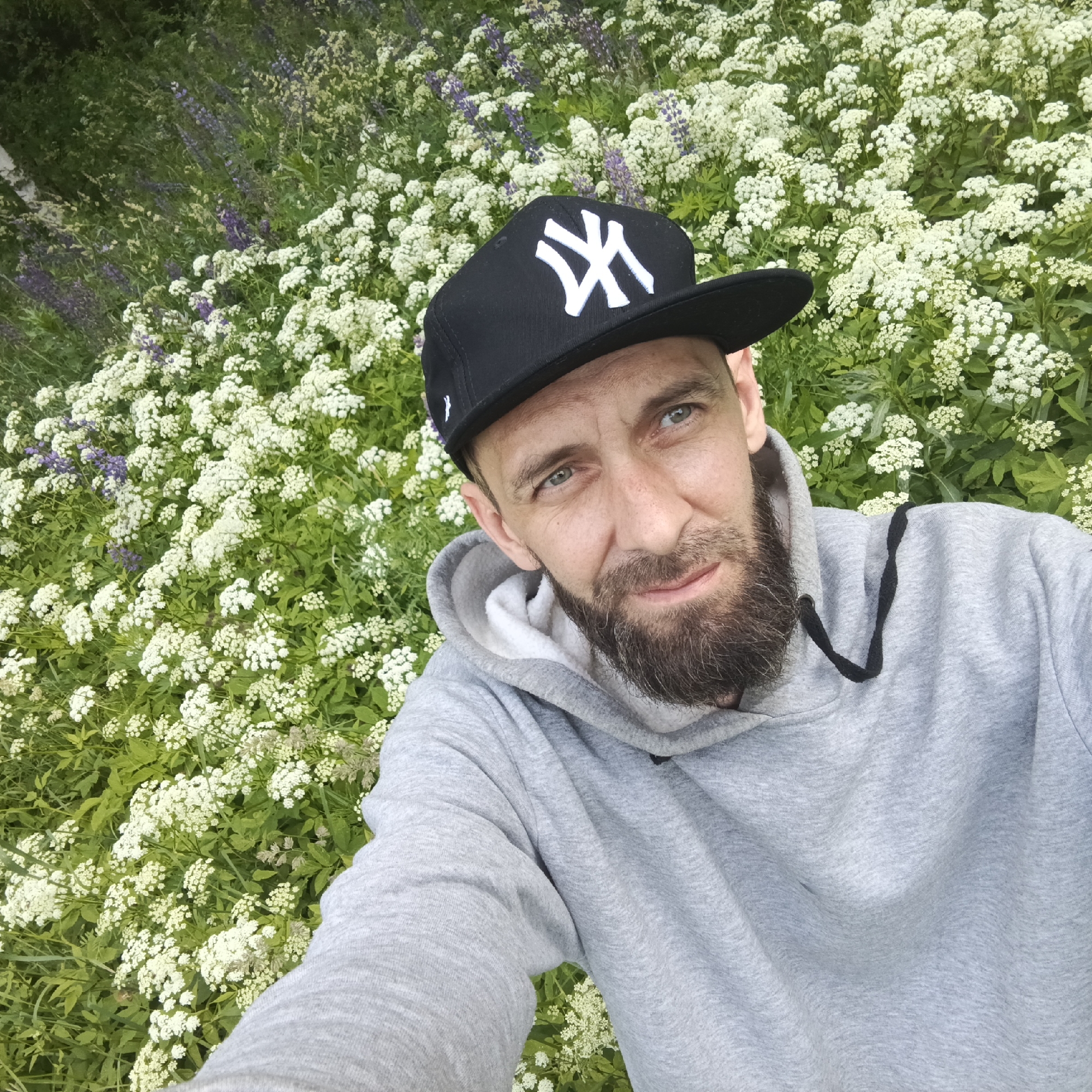 A man with a beard and a New York Yankees baseball cap is taking a selfie in a field of white flowers and green foliage. He is wearing a grey hooded sweatshirt. The background consists of dense green vegetation dotted with clusters of small white flowers and some purple flowers. The lighting suggests it is daytime outdoors. The specific location within the city of Марковский, Russia cannot be determined from the image.