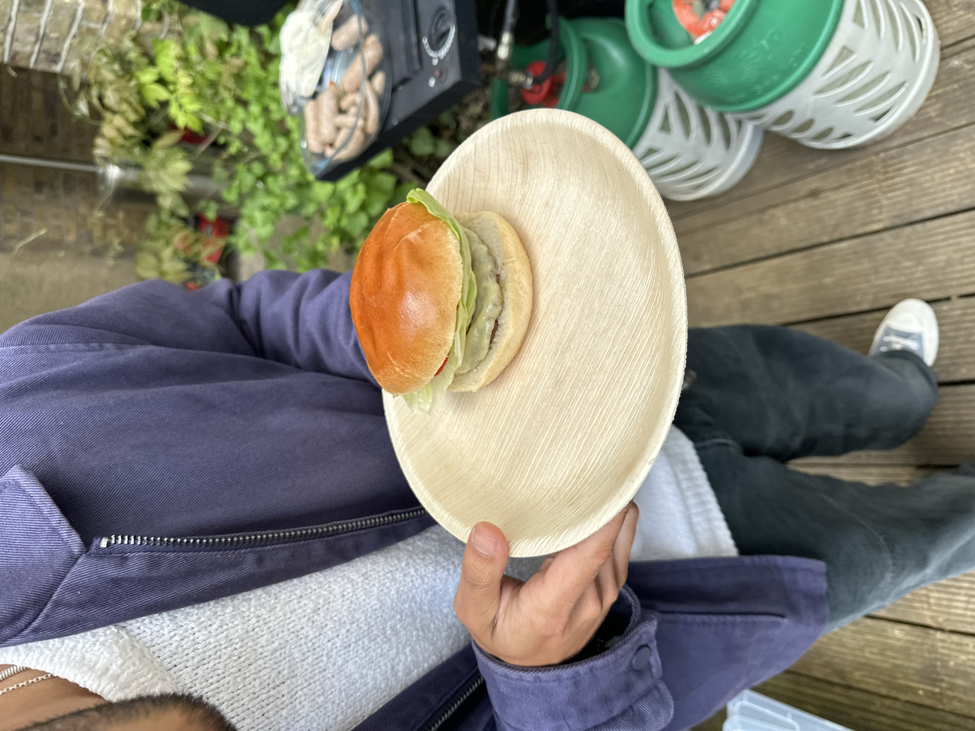 A person is holding a tan palm leaf plate with a sandwich on it. The person is wearing a blue jacket, a gray sweater, and black jeans. The person is also wearing a white shoe with a blue logo. The plate is in focus, and the background is blurred. The background includes a wooden deck, a green gas tank, a silver grill, and some green plants. The sandwich has lettuce and what looks like a meat patty.
There is no explicit evidence to confirm this photo was taken in London, United Kingdom.
