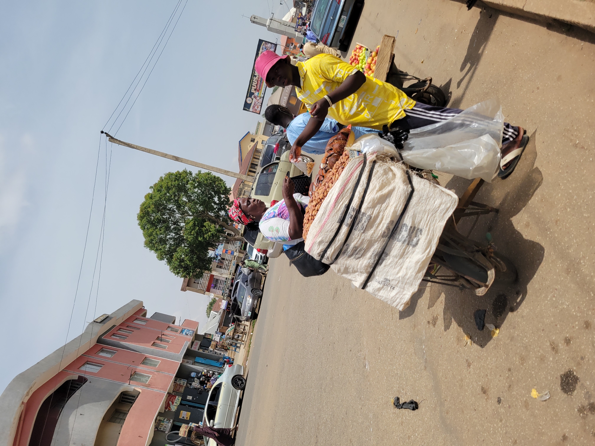 The image shows a street scene in Jos, Nigeria. Two vendors are visible. One, wearing a pink hat and yellow shirt, is selling fruits and nuts from a hand-pushed cart to a woman in a patterned headscarf. The woman is looking up at the vendor and appears to be making a purchase. The cart is overloaded with goods, including a white bag with black stripes. In the background, there are buildings, cars, and other pedestrians. The road appears to be paved. The weather looks sunny.
