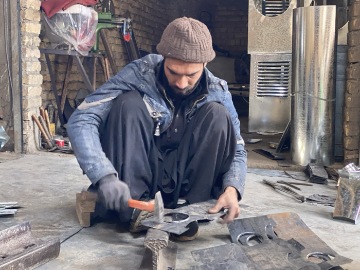 The image displays a male individual actively engaged in metalworking within a workshop located in Kandahar, Afghanistan. The man is observed crouching on a concrete floor, dressed in a brown knitted beanie, a blue denim jacket featuring a white logo on the right sleeve, and dark-colored trousers. His left hand, covered by a black glove, firmly holds a circular piece of sheet metal placed on a wooden block. His right hand, ungloved, wields an orange-handled hammer, which he is using to strike the metal.
Scattered across the concrete floor in the foreground are numerous pieces of cut sheet metal, some exhibiting irregular shapes and circular cutouts. A heavy, dark metal object, possibly an anvil or mold, is visible on the floor in the lower left.
The background reveals structural elements of the workshop, including exposed brick walls. Against the left wall, several tools, resembling axes or mallets, lean vertically. Further back on the left, a green industrial machine with a red wheel is partially visible. Against the right wall, several large, unfinished metal components stand upright; these include two rectangular sections with horizontal louvers, characteristic of evaporative cooler or HVAC panels, and a tall, cylindrical piece of sheet metal. Additional metal scraps and components are visible on the floor and against the walls in the midground and background, indicating an active manufacturing environment.