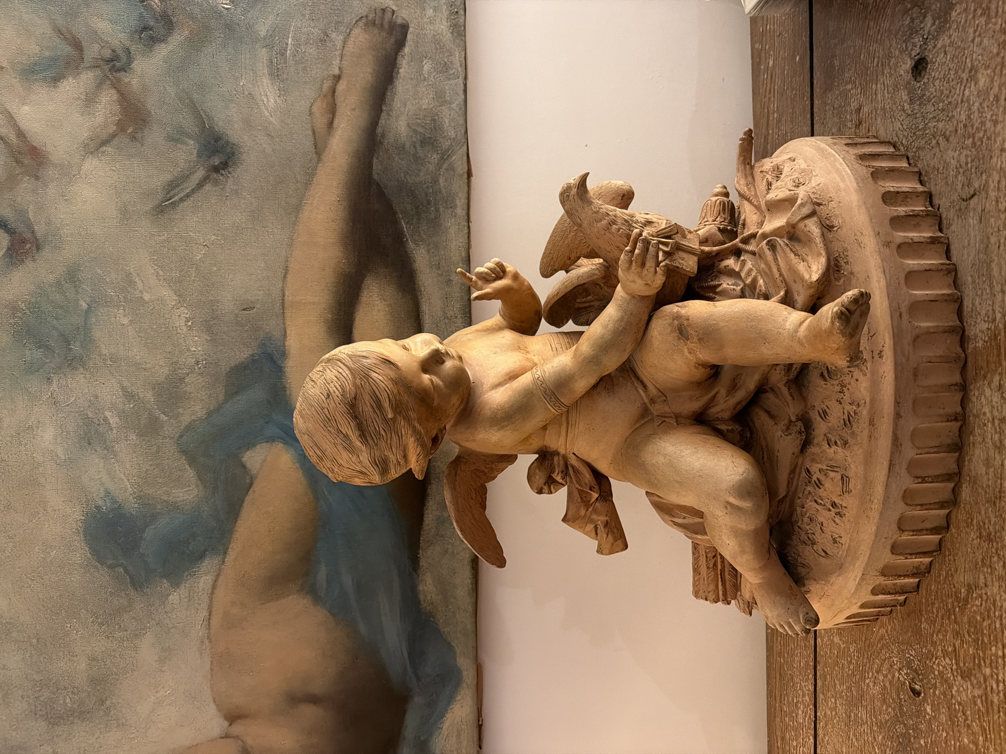 A terracotta-colored statue of a winged cherub occupies the center-right foreground, positioned on a circular, fluted base resting on a rustic wooden table or shelf. The cherub, with short hair and a sash draped around its lower body, holds a small bird in its left hand while its right index finger points skyward. The cherub's gaze is directed towards the bird. Behind the statue, an off-white wall rises, topped by a large, unframed canvas painting. This painting depicts a reclining nude figure, rendered in muted flesh tones and blue drapery, against a light blue and grey background that contains abstract forms resembling flying birds. The overall environment suggests an interior setting, but no specific landmarks are present to determine a precise location within the city of Paris, France.