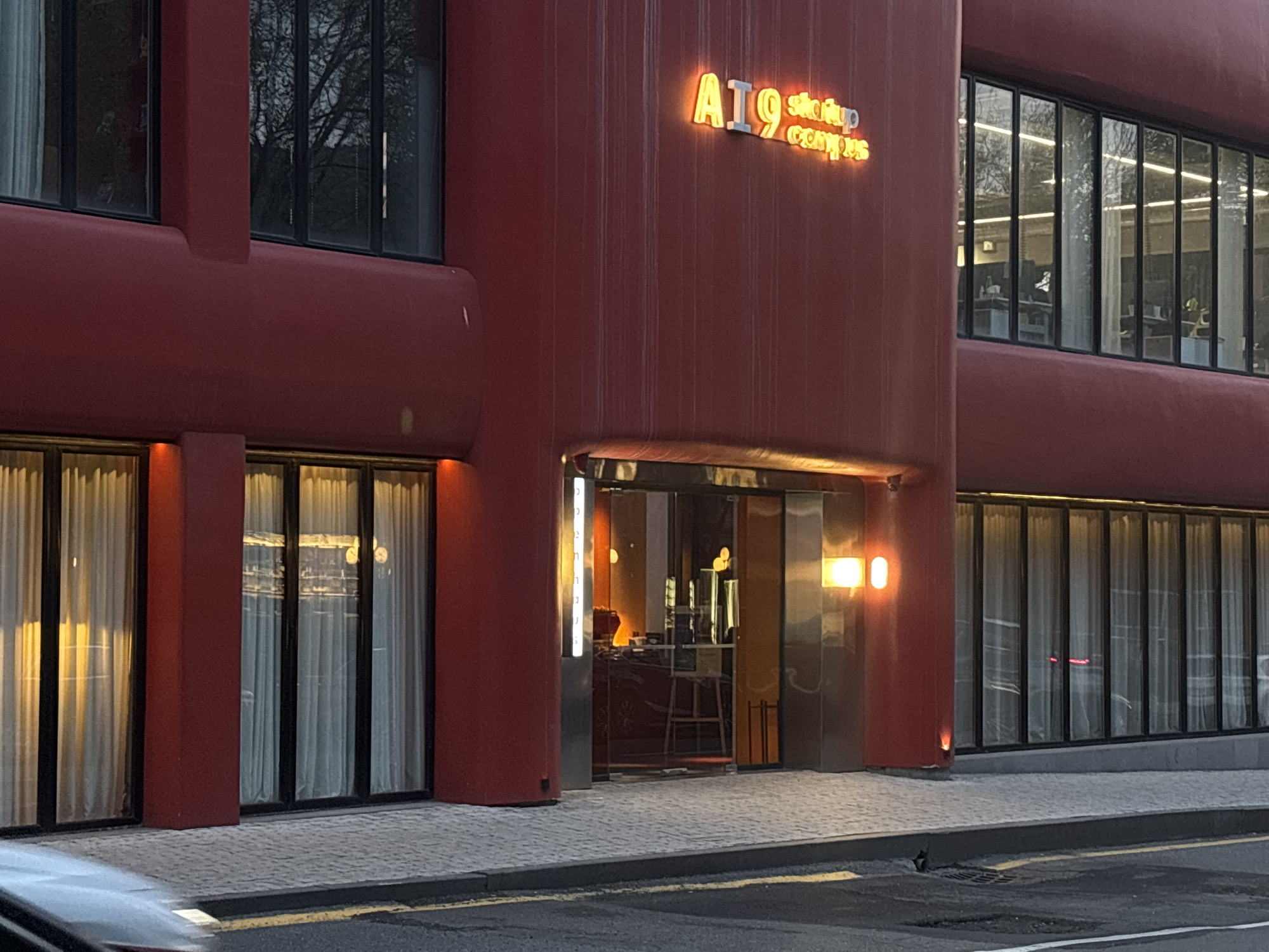 The image shows a reddish-brown building with large windows and a sign that reads "A19 startup campus". The building appears to be located on a street with a paved sidewalk and an asphalt road. The windows have white curtains inside. The entrance has glass doors and metallic framing. Inside, there are tables and chairs. Based on the sign, this is the A19 Startup Campus. While it's difficult to definitively pinpoint the location to Yerevan without further evidence, there is an A19 Startup Campus located in Yerevan, Armenia, which makes it a likely location. No people are clearly visible outside the building, but there appear to be people inside. The overall environment seems urban.