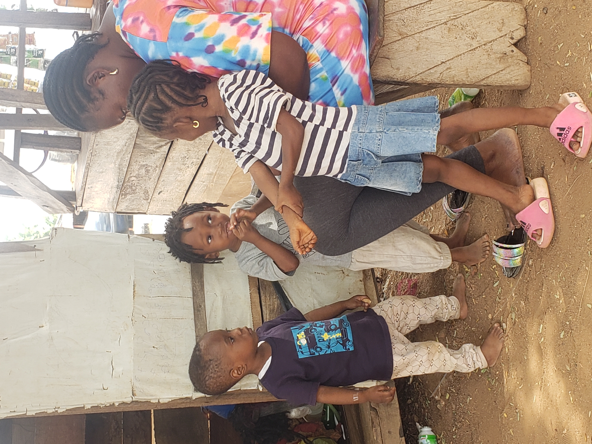 This image, taken in Adankolo, Nigeria, depicts an adult female with three children in a casual outdoor setting. The photograph is oriented with the subjects lying on their sides, suggesting a 90-degree counter-clockwise rotation from the original capture.
The adult female, with dark skin and braided hair, wears a vibrant, multi-colored tie-dye patterned top or dress. She is seated on a sturdy, rough-hewn wooden bench or platform, with her arm around or holding the eldest child. This child, an older girl with braided hair and a small gold hoop earring, is dressed in a black and white striped long-sleeve top and a light blue denim overall skirt. Her feet are adorned with pink Adidas slides. She has one hand holding the arm of the middle child.
The middle child, a boy with dark skin and dreadlocks or braided hair, is seated, wearing a grey t-shirt and light-colored pants, and has one finger near his mouth. His feet wear colorful beaded slides. The youngest child, a boy with short hair, stands barefoot on the dirt ground, dressed in a navy blue t-shirt featuring a blue graphic design and light-colored patterned pants. A small band-aid is visible on his left arm.
The background shows simple wooden structures, including a wooden wall or stall, and a section covered with white corrugated material. This white surface bears some handwritten text and numbers, such as "5 10 10" and "4 4 4", along with other indistinct scribbles. The ground is bare earth. A partially visible green bottle lies on the ground.
The scene conveys an everyday moment of interaction, likely within a family or community context, under bright daytime light. The visible text includes the "Adidas" logo on the older girl's slides and the handwritten numbers on the background structure.