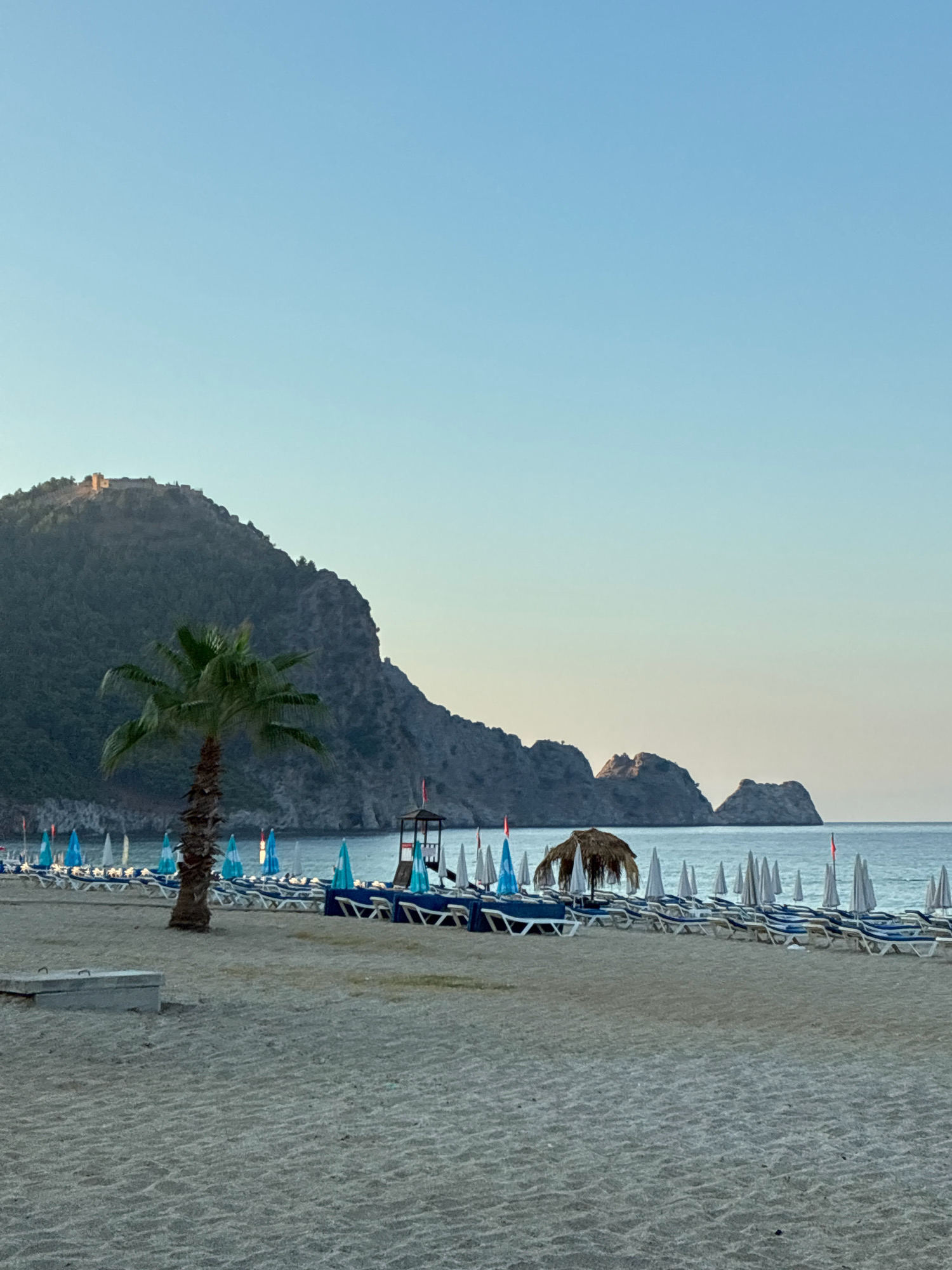 A serene beach scene in Alanya, Turkey, is captured under a clear, pale blue sky. The foreground is dominated by fine, pale sand, marked with subtle ripples from the tide. A single, mature palm tree stands to the left, its fronds rustling gently, providing a tropical accent.
Further into the scene, rows of empty lounge chairs and closed umbrellas are neatly arranged on the sand, suggesting a peaceful, early morning or late afternoon setting. A few of the umbrellas are open, showcasing a light blue and white pattern. A wooden lifeguard tower, equipped with a bench and roof, stands prominently in the middle ground. Beyond the beach, the calm, azure sea stretches to the horizon.
In the background, a dramatic mountain range rises, its slopes covered in dark vegetation. A historic fortress or castle is visible perched on the crest of the largest peak, hinting at the rich history of the region. Further out to sea, jagged rock formations emerge from the water. The overall impression is one of tranquility and natural beauty, characteristic of Alanya's coastal landscape.