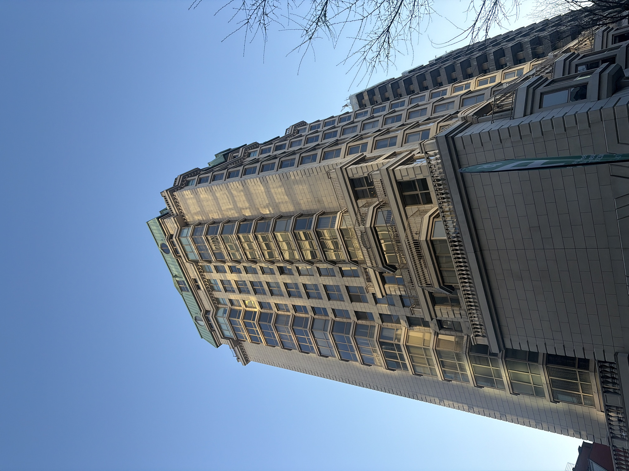 The image presents a tall, modern building seen from a low, upward-looking angle against a bright, clear blue sky in Montreal, Canada. The photograph is rotated, making the building appear diagonally across the frame, extending from the lower left to the upper right.
The building exhibits a distinctive stepped or terraced architectural style, with multiple setbacks creating a layered effect as it rises. Its facade is composed of numerous windows, many of which are angled or project outwards, giving the building a textured and geometric appearance. The exterior material appears to be a light gray or beige, with some upper sections featuring green-colored roof elements, possibly made of metal or oxidized copper. The surfaces of the building and its windows reflect the bright sunlight, creating highlights and shadows that emphasize its intricate design. Decorative railings and patterned panels are visible on various levels, adding to the architectural detail.
Bare tree branches, without leaves, are visible in the upper parts of the frame, suggesting the photo was taken during late autumn, winter, or early spring. The clear, vibrant blue sky indicates fair weather and a sunny day, likely during the late morning or early afternoon given the angle of the light. No people are visible in the scene.
A partially visible green and white sign or banner is attached to the side of the building on the right-hand side. While its colors and general shape are discernible, any specific text or detailed graphic on it is obscured or too blurry to be read. The overall impression is a dynamic shot showcasing the intricate architecture of an urban high-rise under a clear sky.