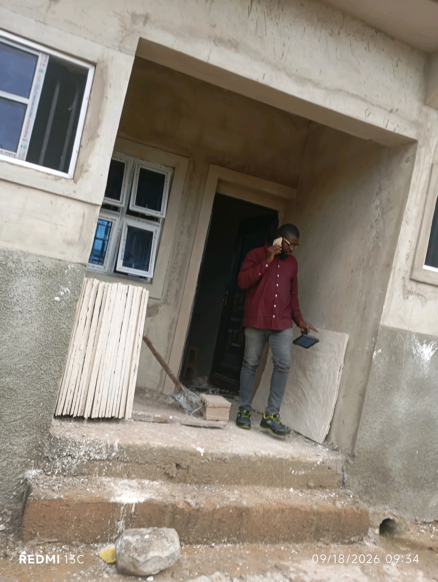A man in a maroon shirt and grey pants is standing on the steps of a building under construction. He is on the phone and holding a tablet. To his left are stacks of wooden planks and a broom. The building appears to be made of concrete with unfinished windows. The scene is outdoors, with debris and construction materials scattered around. The timestamp indicates the image was taken on September 18, 2026, at 9:34 AM. The location is identified as Shendam, Nigeria.