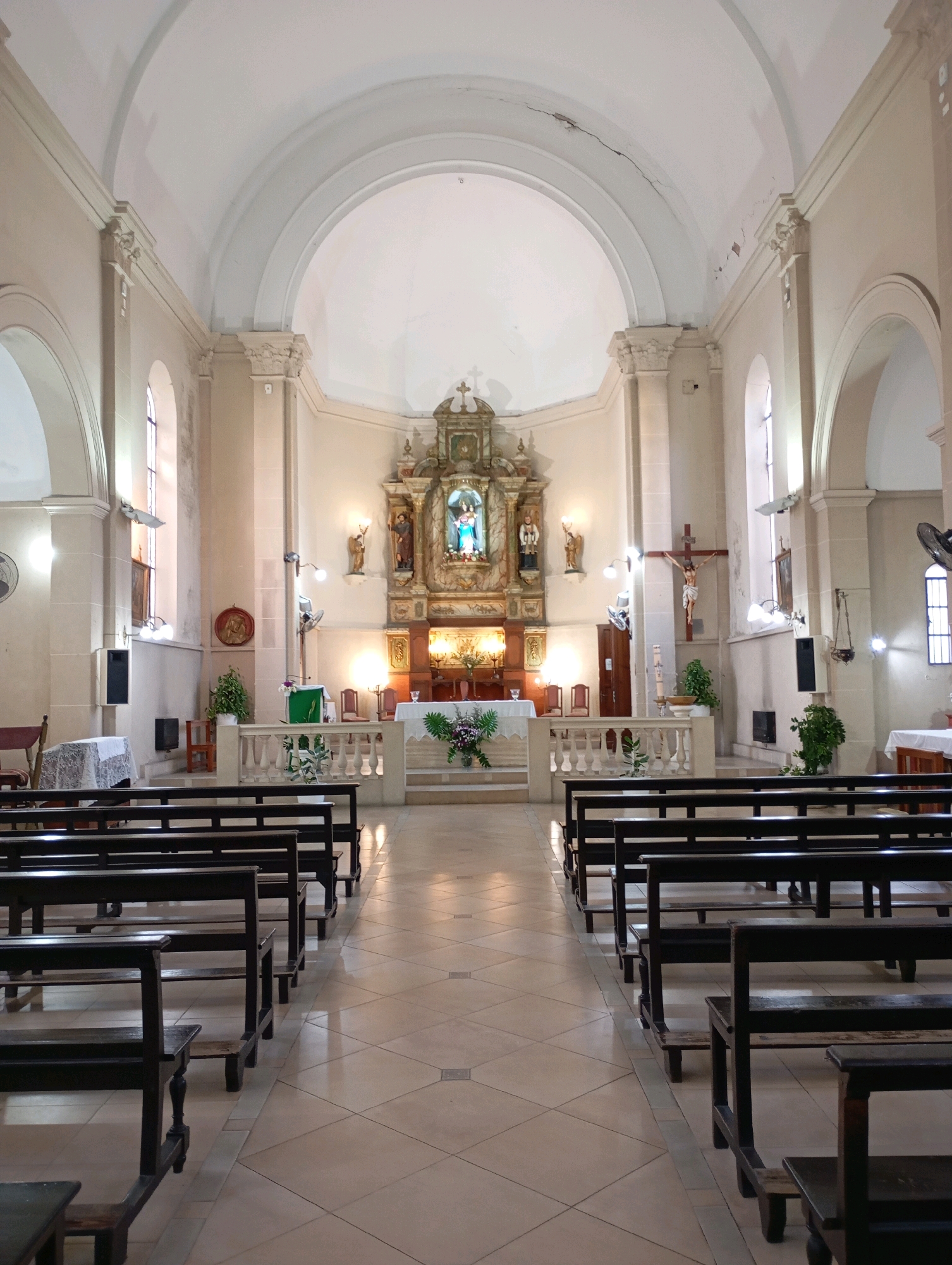 The image depicts the interior of a church, likely located in Berisso, Argentina, given the context. The scene is an interior view facing the altar. The church features a nave with rows of dark wooden pews on either side, separated by a central aisle paved with light-colored square tiles arranged in a diamond pattern. The walls are a light cream color, and the ceiling is vaulted with white plaster, showing some cracks in the upper sections.
At the front of the church, the altar area is prominent. It is adorned with a large, ornate altarpiece featuring religious figures, including a statue of the Virgin Mary in blue. To the right of the altar, a crucifix with Jesus is displayed on the wall. The altar itself is decorated with flowers and lit candles.
Architectural elements include arched windows on the sides, pilasters, and decorative moldings. There are also several light fixtures, fans, and some decorative plants placed strategically throughout the space. The overall impression is one of solemnity and quietude. No people are visible in the image.