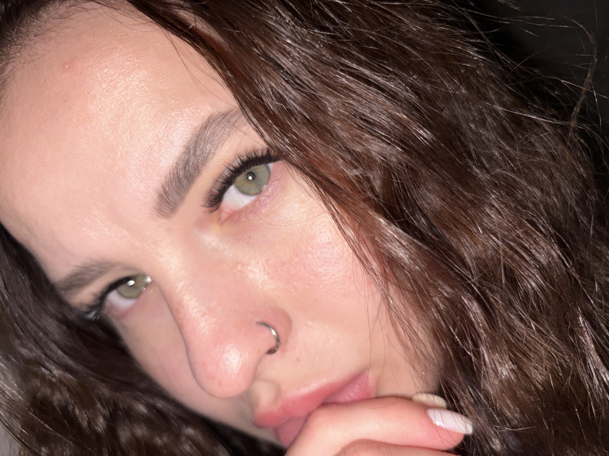Close-up portrait of a young woman with light skin, brown wavy hair, and green eyes. She has long eyelashes and a silver nose ring. Her hand with a white-painted fingernail is near her mouth. The lighting is dim. The image provides no information to identify its location as Krasnodar, Russia, or any other location.