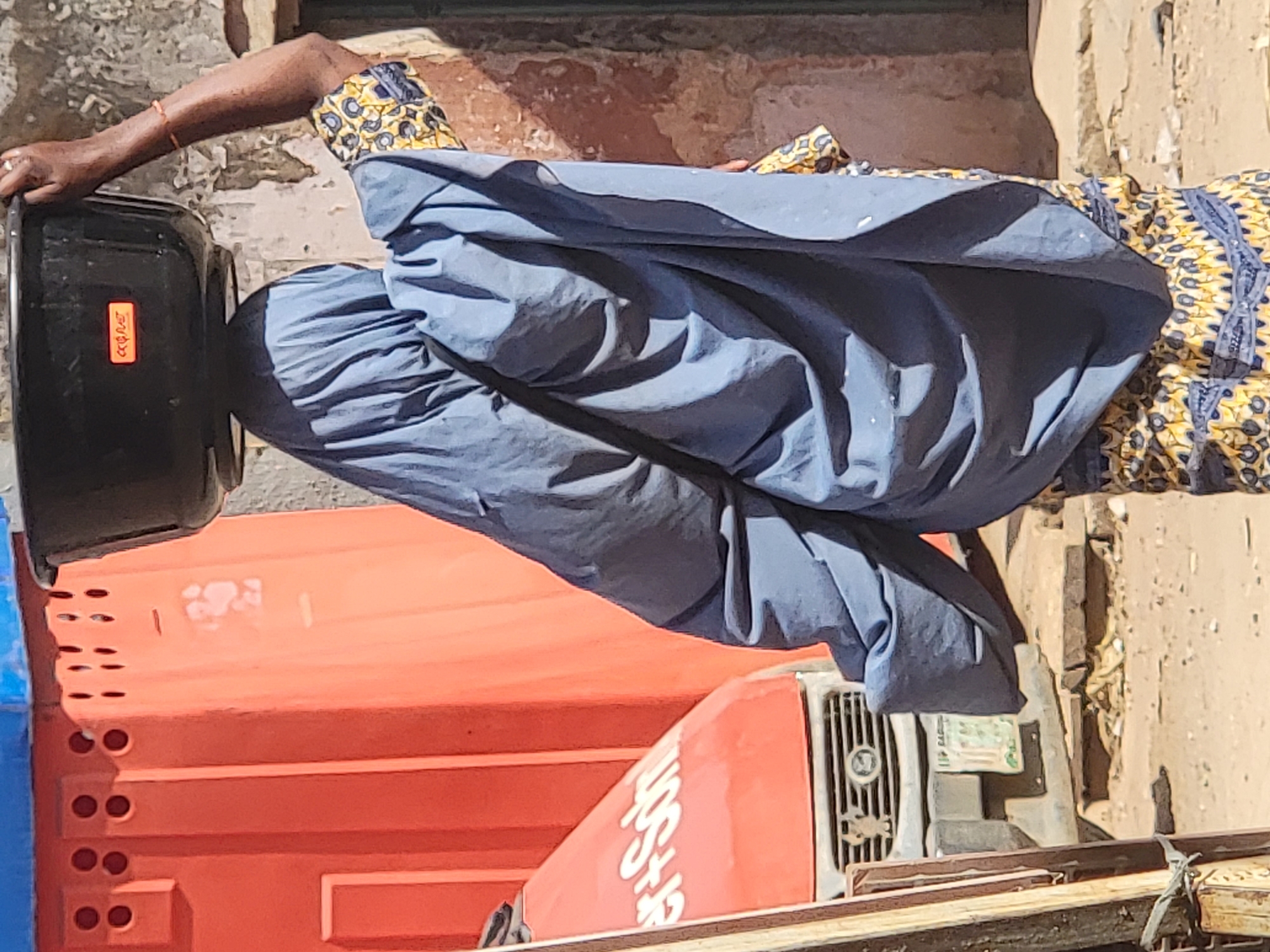 A person wearing a blue gown and patterned yellow and blue sleeves is holding a black bucket in front of an orange truck in Maiburiji, Nigeria. The scene takes place outside, likely on a street or roadside, given the presence of the truck and building materials.
