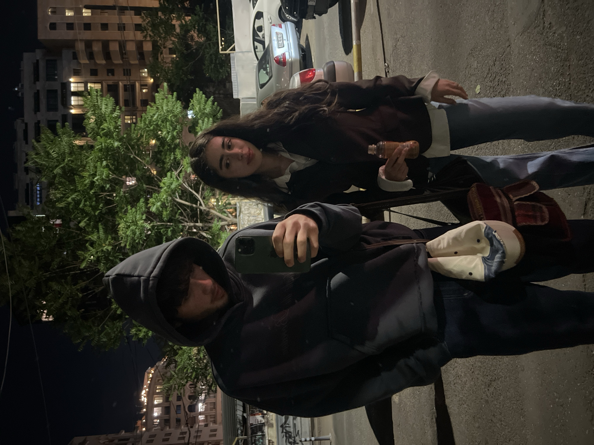 Two people are posing for a photo outdoors at night in Yerevan, Armenia. One person, wearing a dark hoodie and jeans, is holding a smartphone and taking the picture. The other person, positioned slightly behind and to the side, is holding a small bottle. She is wearing a brown jacket over a white top and blue jeans. A red bag and a white cap are on the ground near the people. Cars are parked in the background. Buildings with illuminated windows and trees are also in the background.
