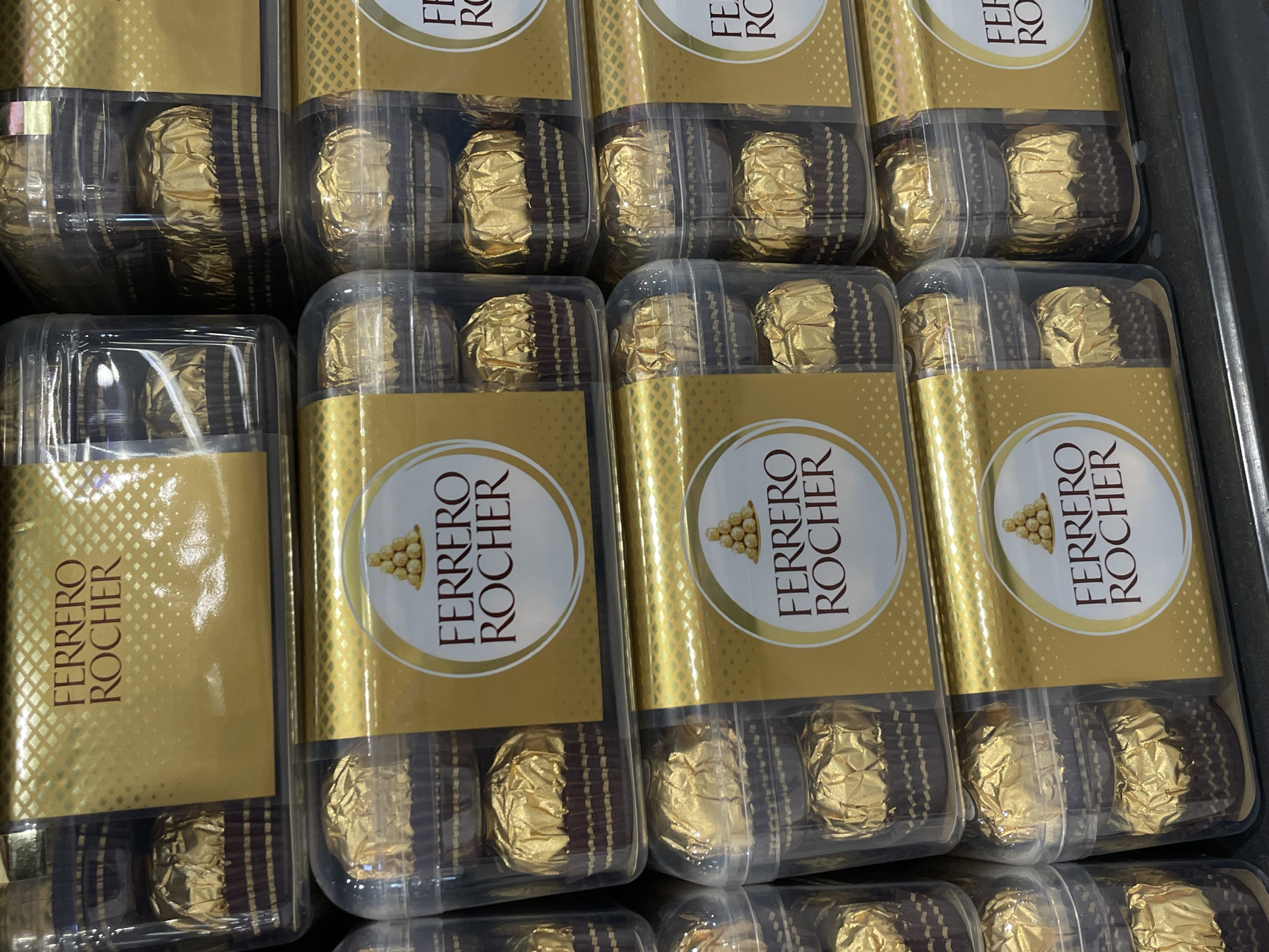 A close-up, eye-level shot shows stacks of Ferrero Rocher chocolates, a popular brand known for its hazelnut and chocolate confectionary. The chocolates are individually wrapped in shiny gold foil and presented in clear plastic packaging. The brand's distinctive gold and brown packaging is visible on the boxes, featuring the "Ferrero Rocher" logo and an image of the chocolates. The arrangement suggests a retail display, likely in a supermarket or a confectionery store. The lighting is bright and even, highlighting the reflective surfaces of the chocolate wrappers and packaging. There are no people visible in the image, and the focus is entirely on the product. The scene is clean and organized, indicative of a commercial setting. The stacked nature of the boxes implies a large quantity of the product, ready for sale. The overall impression is one of abundance and premium presentation. This image could have been taken at a store within Davao City, Philippines, given the popularity of such imported chocolates in the region.