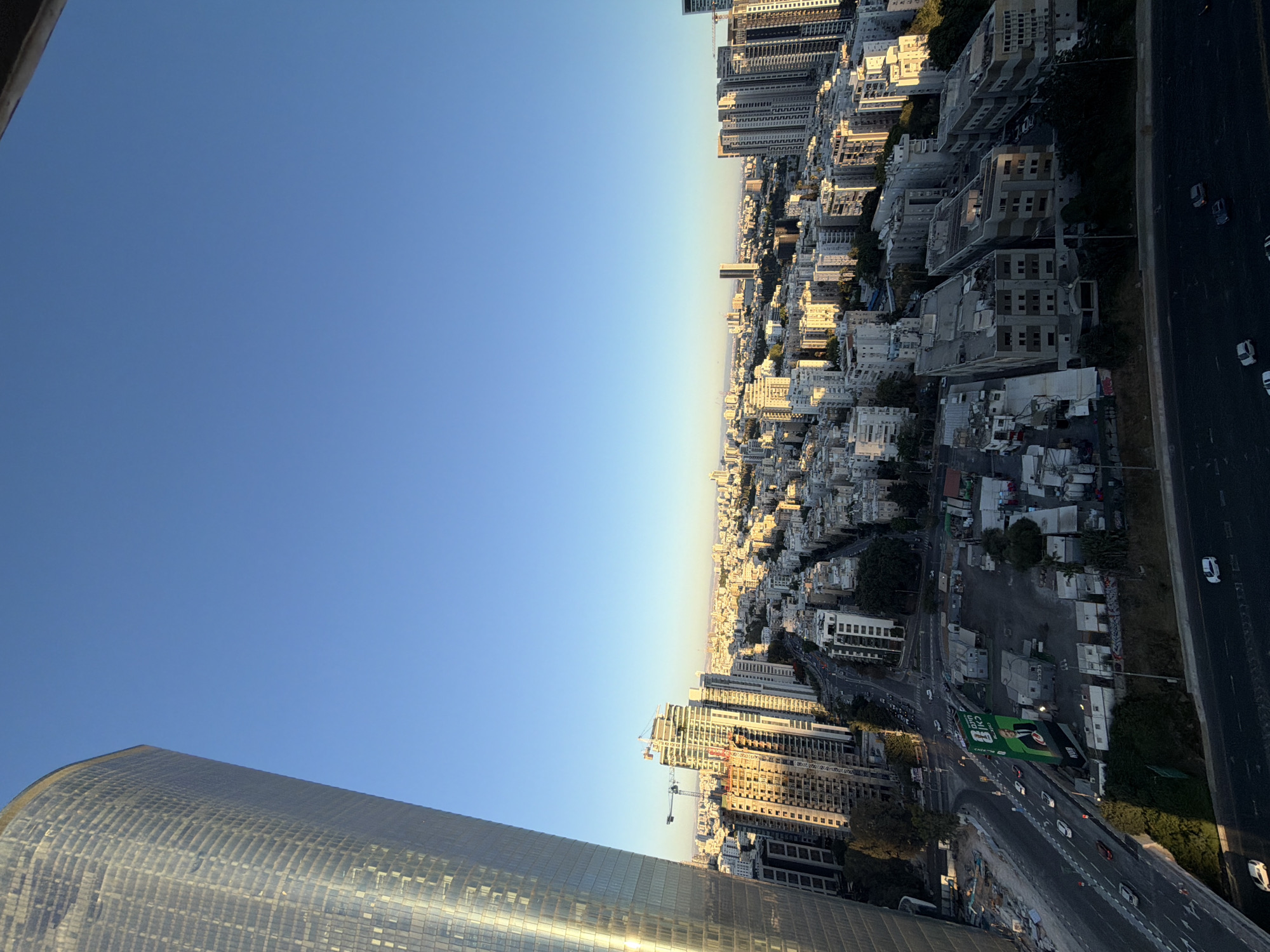 This is an aerial, eye-level, wide shot taken from a high vantage point, looking down and across a dense urban cityscape. A tall, modern glass-covered skyscraper dominates the left side of the frame, reflecting the clear blue sky and the warm, low-angled sunlight, suggesting either sunrise or sunset. The skyscraper's curved facade is textured with a grid pattern of windows.
Beyond the skyscraper, the city stretches out, a panorama of mid-rise and high-rise buildings. Many of these structures are residential or office buildings, characterized by their light-colored facades and balconies, typical of Tel Aviv's architecture. The sunlight casts long shadows across the buildings, highlighting the contours of the city.
In the middle ground, a multi-lane road system is visible, with cars driving along. There's evidence of ongoing construction, with cranes and partially built structures interspersed among the completed buildings. A large, green billboard featuring a man in a suit is prominently displayed near an intersection. The billboard has text in Hebrew.
The environment appears to be a bustling city, likely Tel Aviv, Israel, given the architectural style and the Hebrew text on the billboard. The sky is clear and vast, devoid of clouds. The overall impression is one of a modern, active, and developing urban environment bathed in natural light. No individuals are clearly discernible in the image, but the presence of cars and infrastructure indicates human activity and occupation.