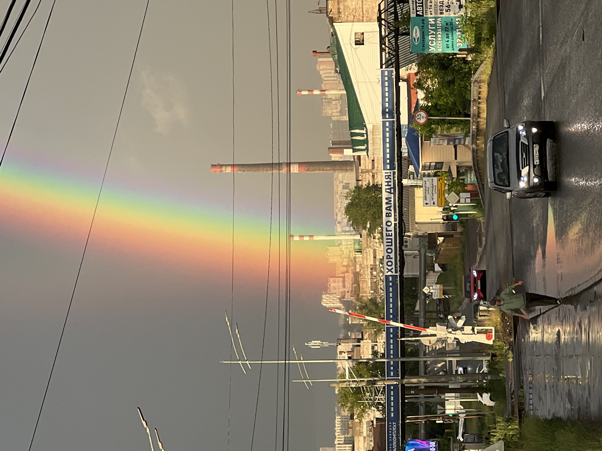 The image depicts a street scene in Izhevsk, Russia, under a dramatic sky illuminated by a vivid rainbow arching across the frame. In the foreground, a man in a green hoodie and dark pants walks away from the camera on a wet, reflective road, possibly after a rainfall. To his right, a silver SUV is driving towards the viewer, its headlights on. Behind them, a red car is seen at a distance on the road.
A railway crossing barrier is visible, with its red and white striped arm raised. Above the crossing, a banner reads "ХОРОШЕГО ВАМ ДНЯ!" (Have a good day!). The background features industrial elements, including tall smokestacks, one of which is red and white striped. Several buildings, some with green roofs, and other urban infrastructure are present. Power lines crisscross the sky. Signs are visible, including one that appears to advertise services and a circular road sign with the number 3.5. The overall impression is of an industrial city on a day with lingering weather effects, where a vibrant natural phenomenon is juxtaposed with urban structures.