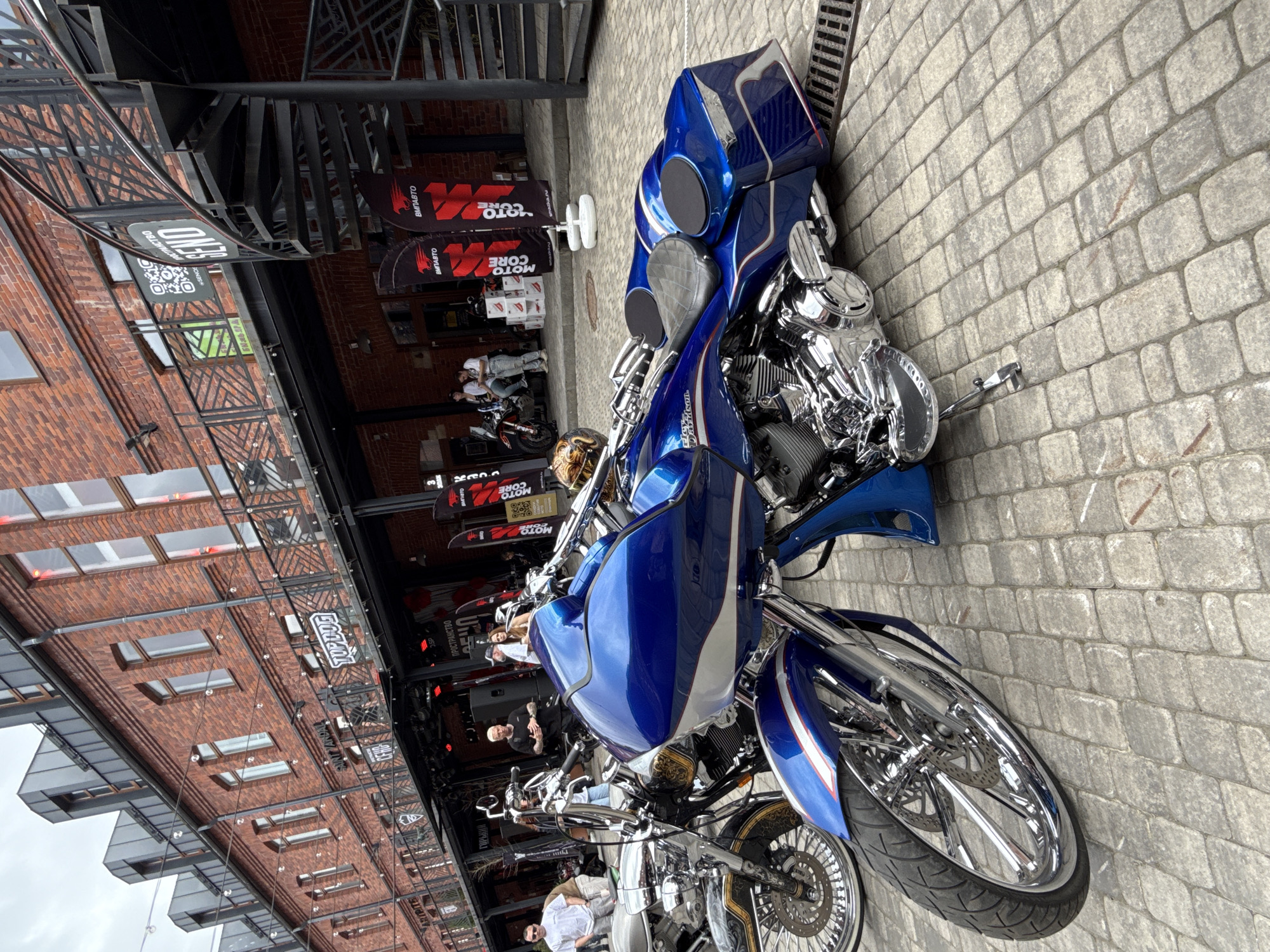 This media file shows a street scene in Saint Petersburg, Russia, featuring several custom motorcycles parked on a cobblestone pavement. The dominant motorcycle is a large blue and silver touring bike with extensive chrome detailing, including its engine and wheels. Another chrome-heavy motorcycle, possibly a cruiser, is partially visible in the foreground.
The scene appears to be part of an outdoor event or gathering, suggested by the presence of promotional banners and signs. Visible text on these banners includes "MOTO CORE," "ВМПАВТО," "TOP DOG," "SAINT VANDAL," and "ГОРЬКИЙ," which are likely related to motorcycle brands, events, or businesses. A QR code is also visible on one of the banners.
Several people are present, some casually dressed and interacting, while others are standing near or looking at the motorcycles. The building in the background is a brick structure with large windows, and a distinctive spiral staircase is visible. The overall atmosphere seems relaxed and indicative of a motorcycle enthusiast event. The lighting suggests it is daytime, with no strong shadows, indicating overcast or diffused light. The presence of branded materials and the concentration of custom bikes point towards a dedicated motorcycle event or exhibition.
