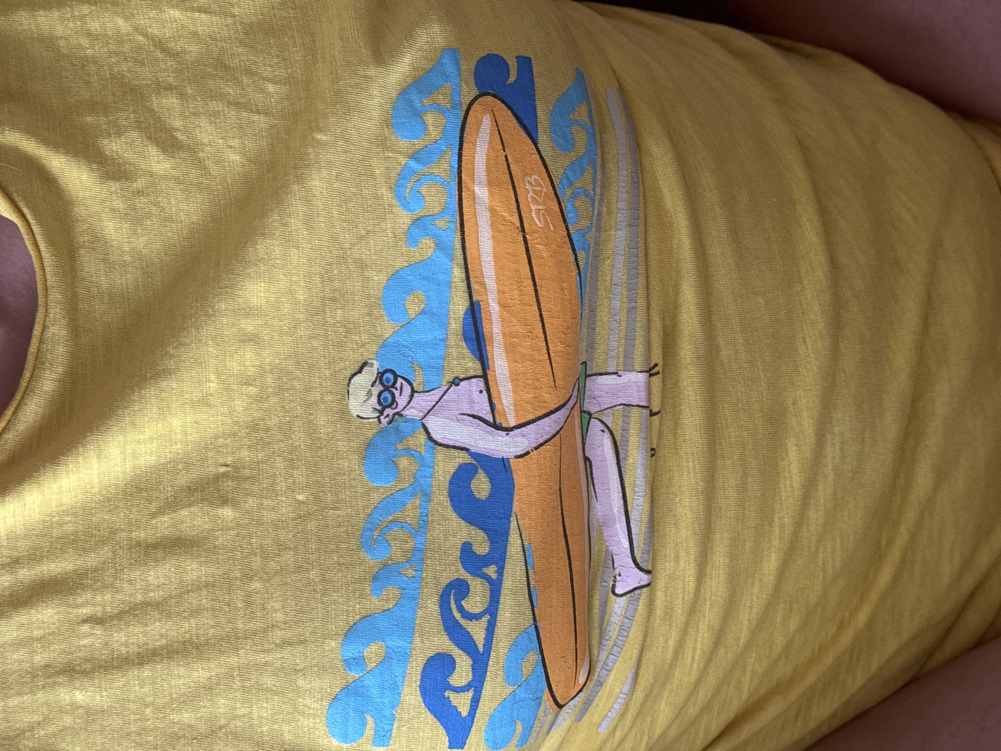 The image shows a yellow t-shirt with a cartoonish design featuring a man hugging a surfboard. The design includes blue wave patterns in the background. The surfboard has the letters "STB" on it.
There is no information in the image to suggest that it is located in the city of Ново-Никольское, Russia.