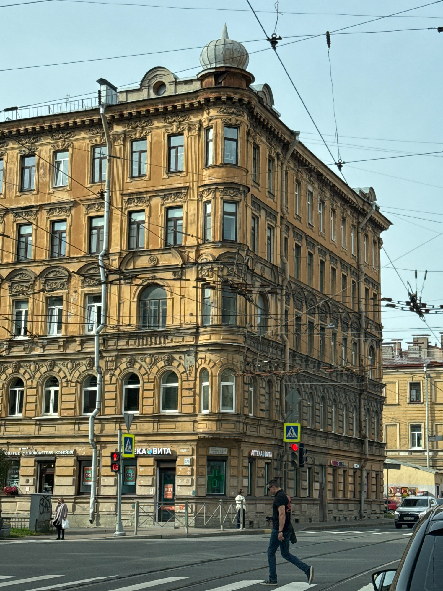 The image shows a street scene in Saint Petersburg, Russia, featuring a large, ornate yellow building with multiple stories and arched windows. The building has intricate architectural details, including decorative moldings and a domed structure on its roof. Overhead, a complex network of wires, likely for trolleybuses or trams, crisscrosses the sky. In the foreground, a pedestrian crossing is visible, with a man walking across the street and a woman standing on the sidewalk. Traffic lights and street signs are present, indicating an intersection. Several businesses are located on the ground floor of the building, including shops with Cyrillic signage, such as "COFFEE MICRO-ROASTERS КОФЕЙНЯ" and "АПТЕКА ВИТА." A white car is parked further down the street, and another vehicle is partially visible in the lower right corner of the image. The sky is overcast, suggesting a cloudy day.