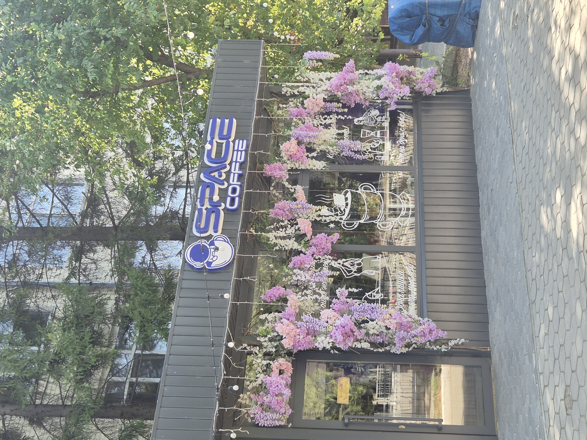 The image shows a storefront for "SPACE COFFEE" in Kokshetau, Kazakhstan. The facade is gray, with a prominent sign featuring the words "SPACE" and "COFFEE" in blue lettering, accompanied by a stylized astronaut logo. The storefront is decorated with hanging floral arrangements in shades of pink and purple, with some white flowers interspersed. The windows of the shop are adorned with white line drawings, including a depiction of a coffee cup with steam, a person doing yoga, and a coffee maker. A yellow sign reading "ЖАБЫК ЗАКРЫТО" (Closed) is visible on the glass door. The area in front of the storefront is paved with hexagonal gray tiles. To the right of the building, a large object covered in a blue tarp, secured by chains, is visible. The scene is set outdoors, with trees and a building visible in the background, suggesting an urban environment.
