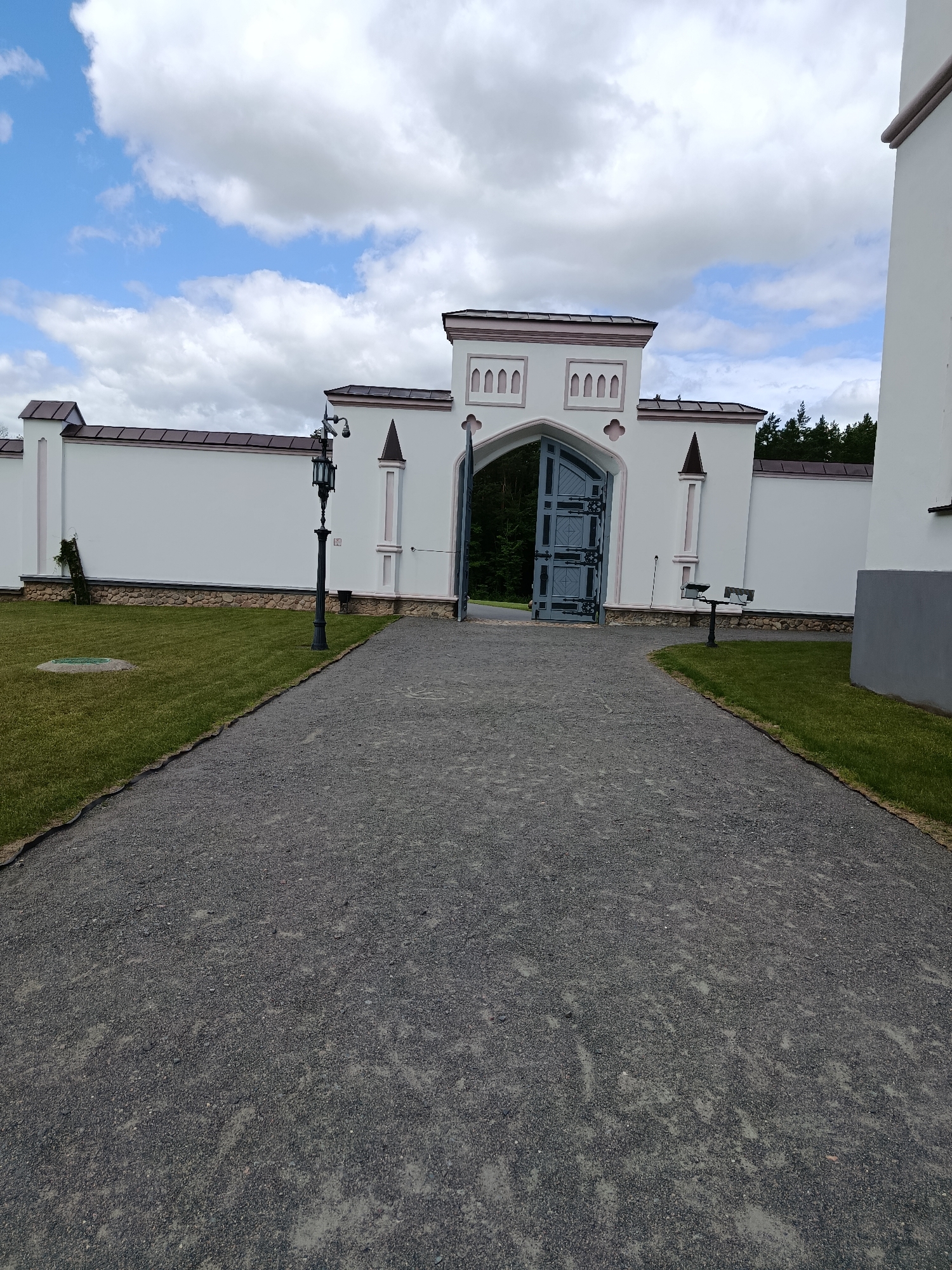 A wide-angle, eye-level shot captures the exterior of the Palace of the Princes of Sapieha in Kosava, Belarus. The foreground is dominated by a grey gravel pathway that curves to the right. To the left of the path is a well-maintained green lawn. The pathway leads towards an open, ornate grey metal gate set within a white archway. The archway is part of a larger white structure with decorative elements. Beyond the gate, a dense forest can be seen. To the left of the pathway and lawn is a white wall with a brown roofline, and a black lamppost stands near the wall. A bench is visible on the grass to the right of the gate. The sky above is a mix of white clouds and blue.