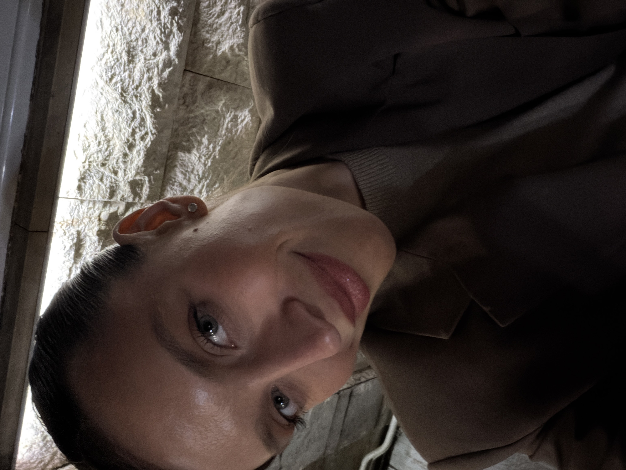 A selfie of a woman with slicked-back dark hair, fair skin, and light-colored eyes. She is wearing a brown blazer over a light beige sweater. The background features a textured wall, possibly stone or stucco, illuminated by a light source. Part of a window frame or other structure is visible to the left. Without additional context, the location in Budva, Montenegro cannot be determined.