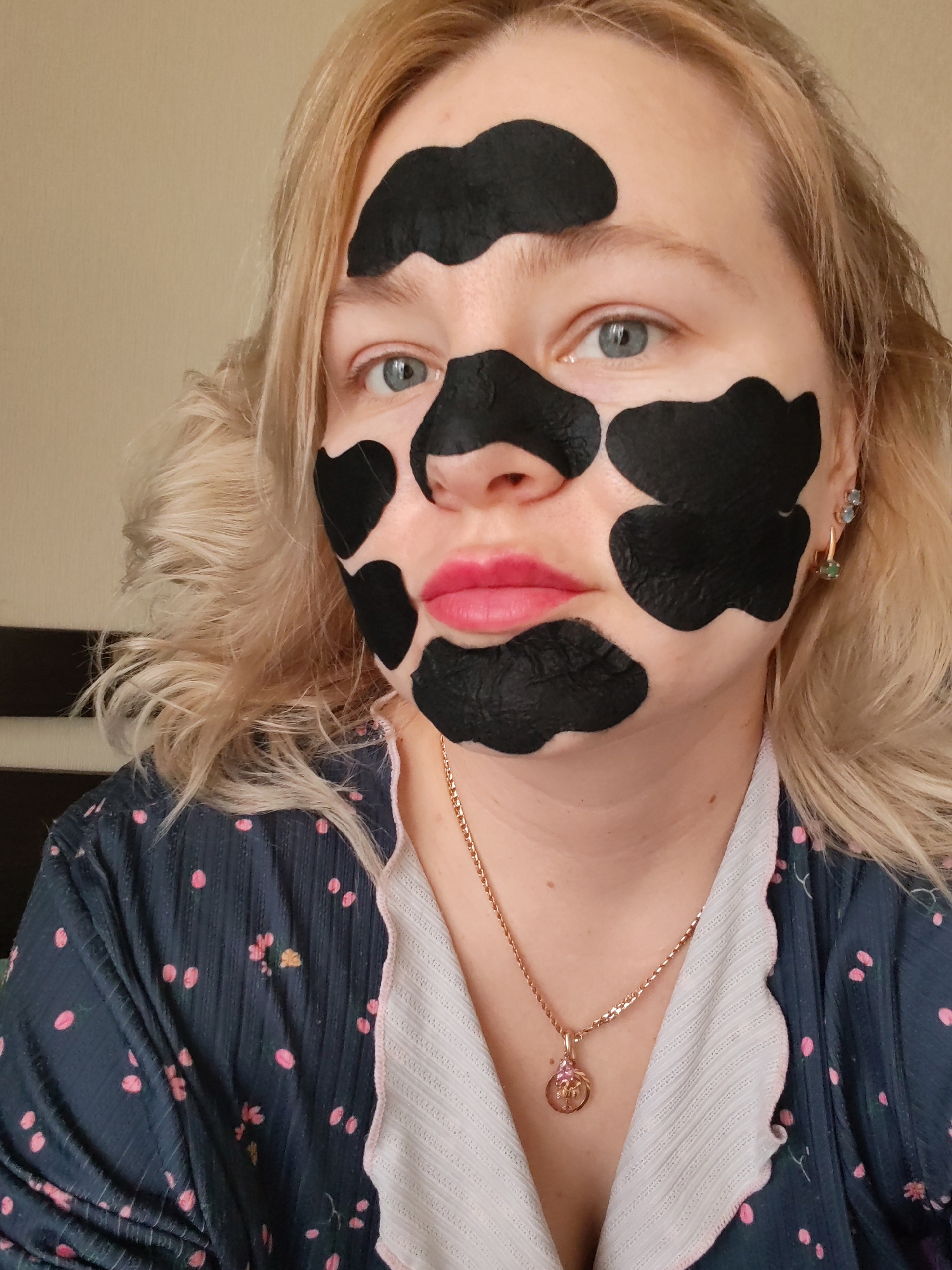 A blonde woman is depicted in a close-up, chest-up shot, engaging in a self-care routine. She has light-colored hair, possibly styled in soft waves, framing her face. Her light blue-grey eyes gaze directly at the viewer, and her lips are painted a vibrant pink.
The most prominent feature is the black peel-off mask applied in irregular, patchy shapes across her face, covering her forehead, cheeks, nose, chin, and above her upper lip. She is wearing a dark blue robe adorned with small pink polka dots and subtle floral patterns, with a white, ribbed lapel edged in pink. A delicate gold-toned chain necklace with a small, ornate pendant featuring pink and possibly other colored stones rests on her chest. In her right ear, she wears two earrings: a small stud with a light blue stone and a drop earring with a green gemstone.
The background is a plain, light beige or tan wall, indicating an indoor setting, likely a home in Izhevsk, Russia. A darker brown object, possibly furniture, is partially visible on the left side of the frame. The lighting is bright and even, suggesting daytime. Her expression is neutral and composed, conveying a sense of calm during her beauty regimen. There is no visible text in the image.