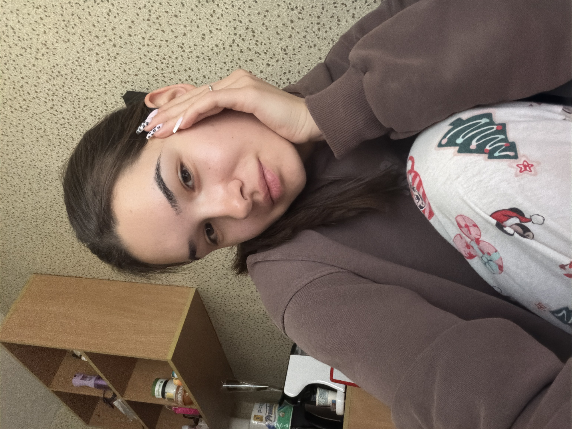 A young woman is captured in a casual selfie, taken indoors in Sofia, Bulgaria. The image is rotated 90 degrees clockwise. She has dark hair, dark, well-defined eyebrows, and light brown eyes, looking directly at the camera with a gentle, relaxed expression and a soft smile. She is wearing a plain brown hoodie or sweatshirt. Her left hand, adorned with a ring on her ring finger, rests casually near her temple. Her long nails feature a stylish design with some having white tips and others a white base with black leopard spots.
She is also wearing white bottoms, likely pajama trousers, decorated with festive, Christmas-themed patterns. These patterns include green Christmas trees, red and white candy canes, and images resembling Minnie Mouse in a Santa hat.
The background consists of a light beige or off-white wall with a subtle, textured pattern. To the woman's left, a wooden shelf unit with multiple compartments is partially visible. Items on the shelves include a purple spray bottle, a white tub labeled "BODY CREAM," and a green-labeled bottle showing partial text like "pick..." and "premium." The scene conveys a comfortable and informal atmosphere, likely within a home.